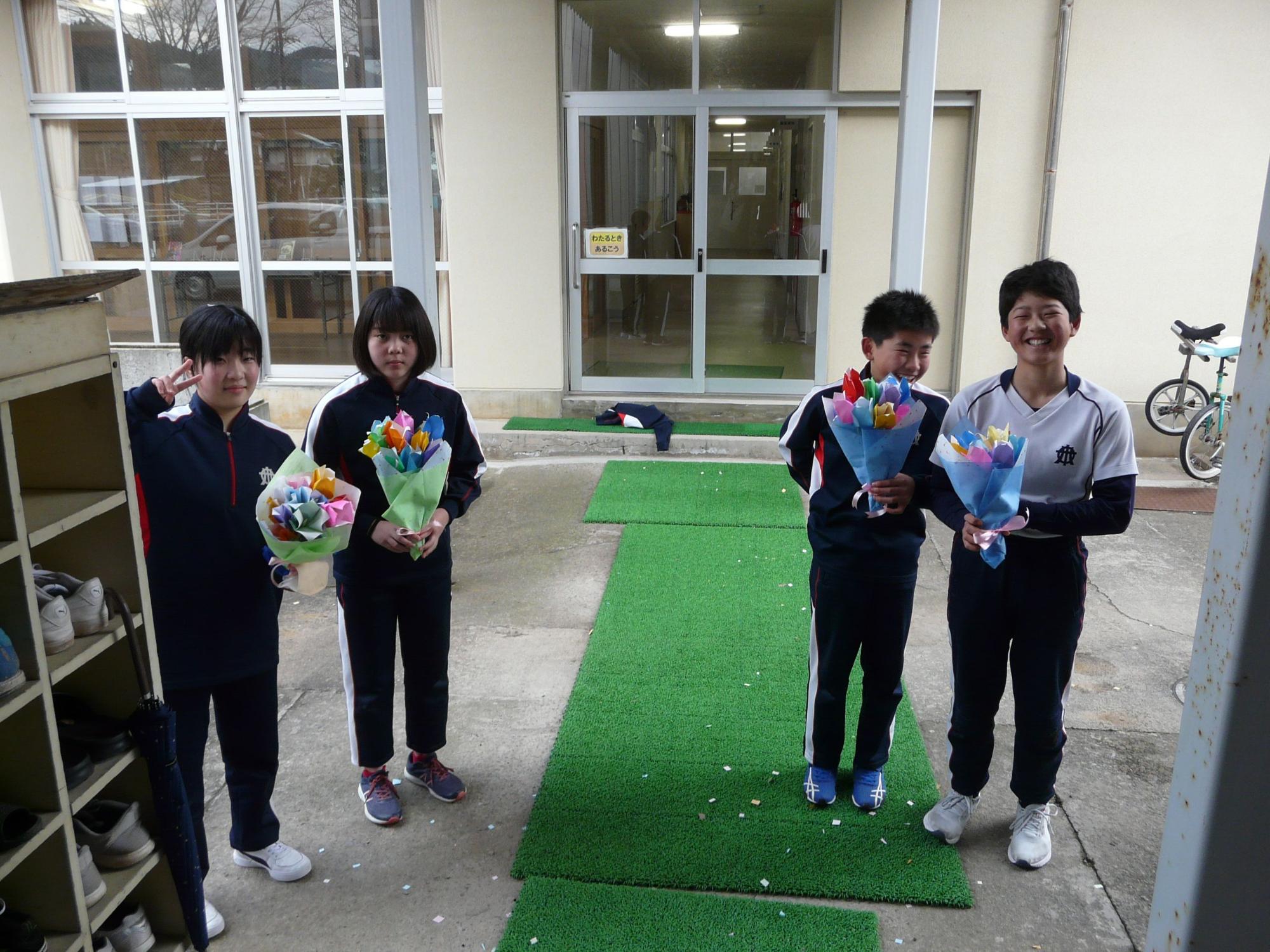 昇降口で花束をもって待っている様子の4人の児童の写真