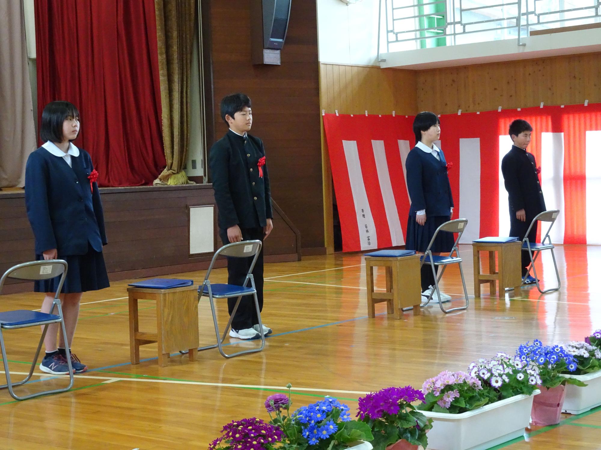 体育館で赤い花を胸元につけた4人の児童が起立している写真