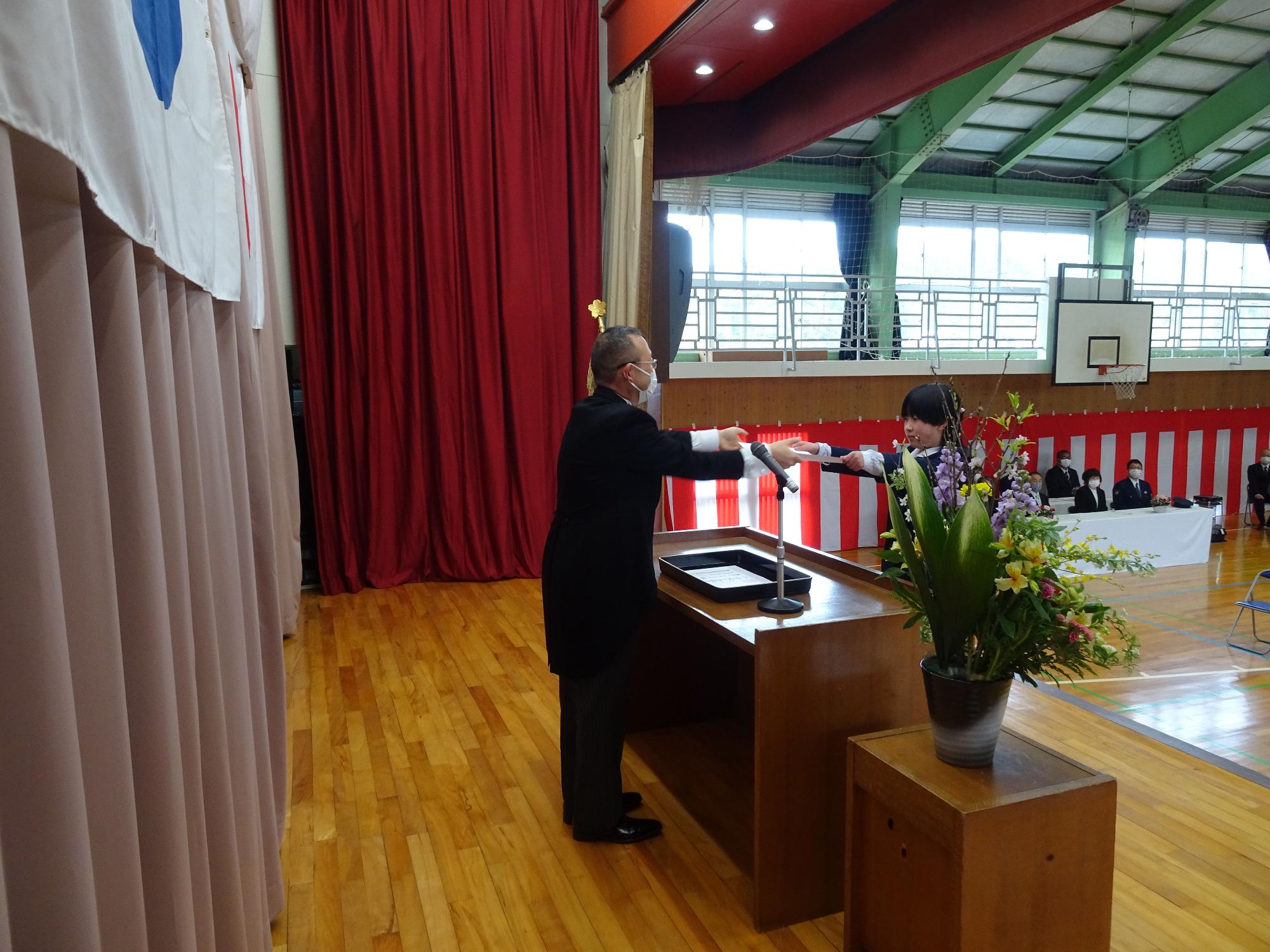 体育館で行われている卒業式で卒業証書を授与されている写真