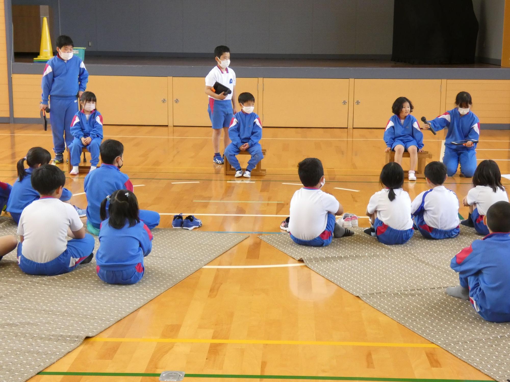 体育館で青いジャージを着た児童たちが座ってレクリエーションをしている写真