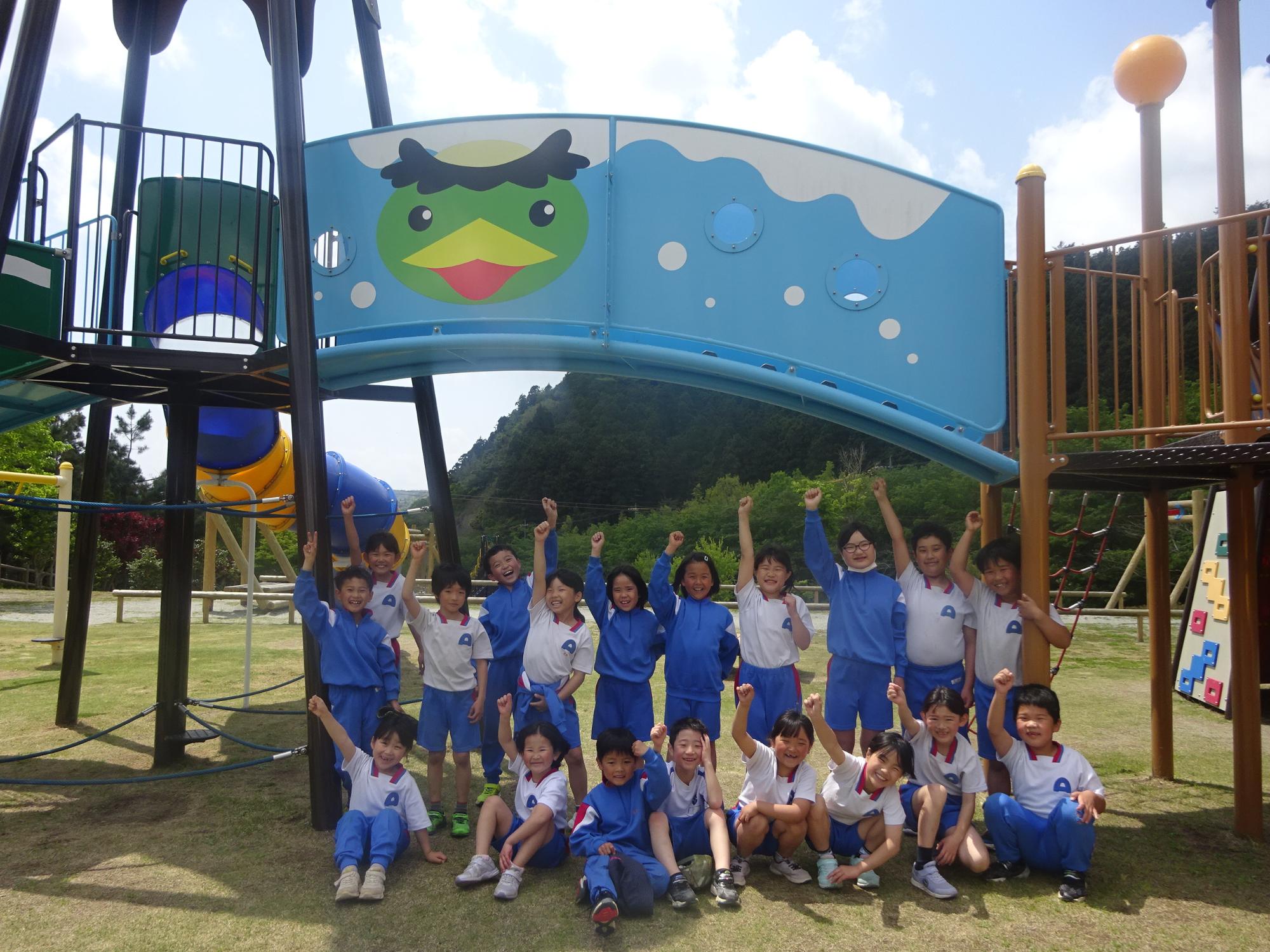 公園で遊具を背景に児童たちが腕を上げて集合写真を撮っている写真