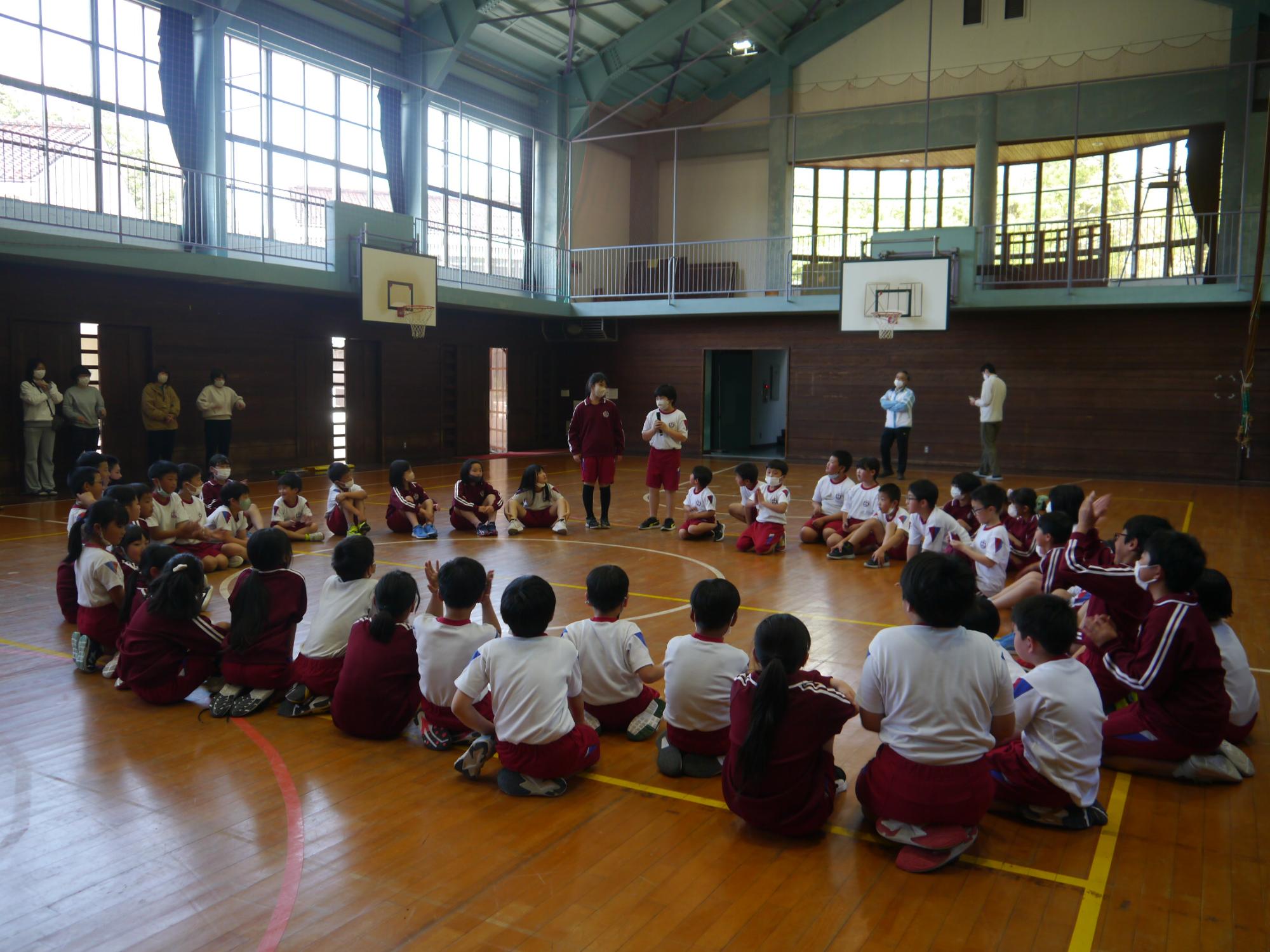 体育館で児童たちが円になって話を聞いている写真