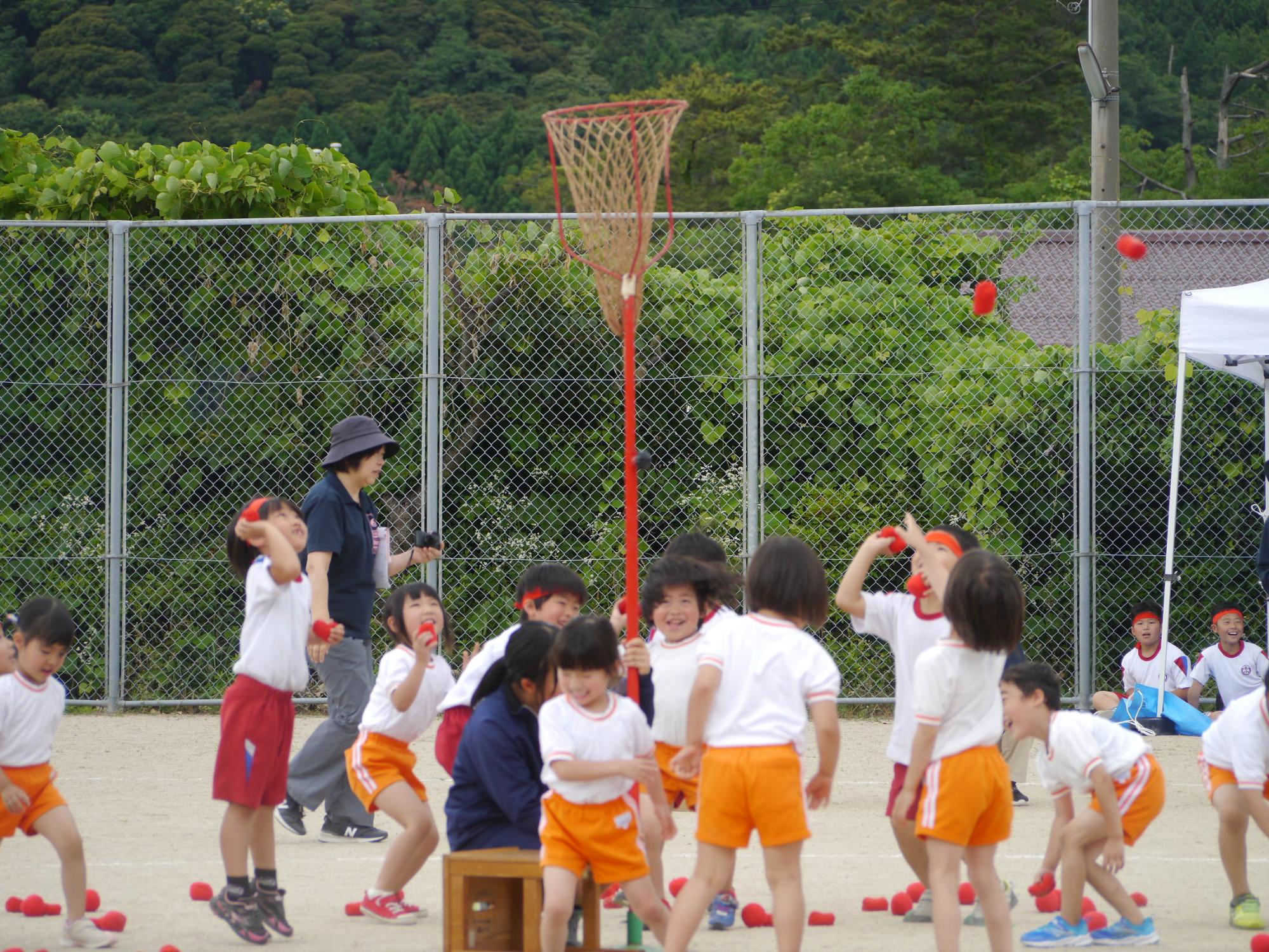 校庭でオレンジ色の体操着を着た児童たちが玉入れをしている写真