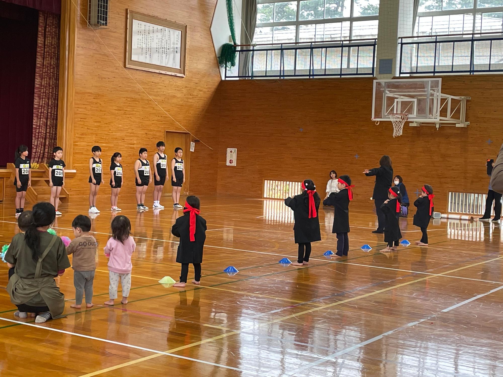 体育館の中で横一列に立って並ぶ選手児童達と、向かい合うように並んで赤い鉢巻きを締めてエールを送る園児達の様子の写真