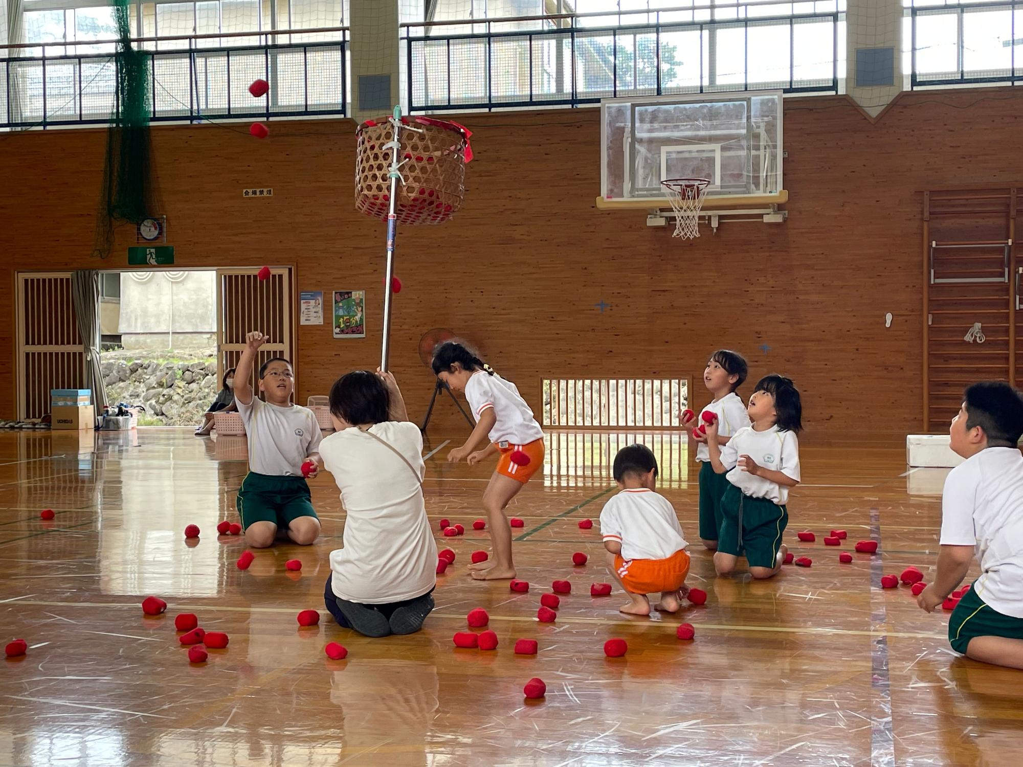 体育館の中で玉入れの網カゴをめがけて赤い球を拾い放り投げる園児と児童たちの写真