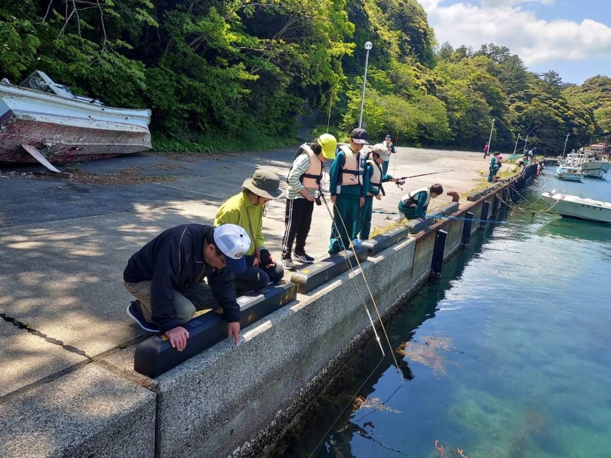 海辺の岸壁の縁に並び釣り竿を手にして魚釣りをしている児童たちと、付き添ってその様子を見守る大人の写真