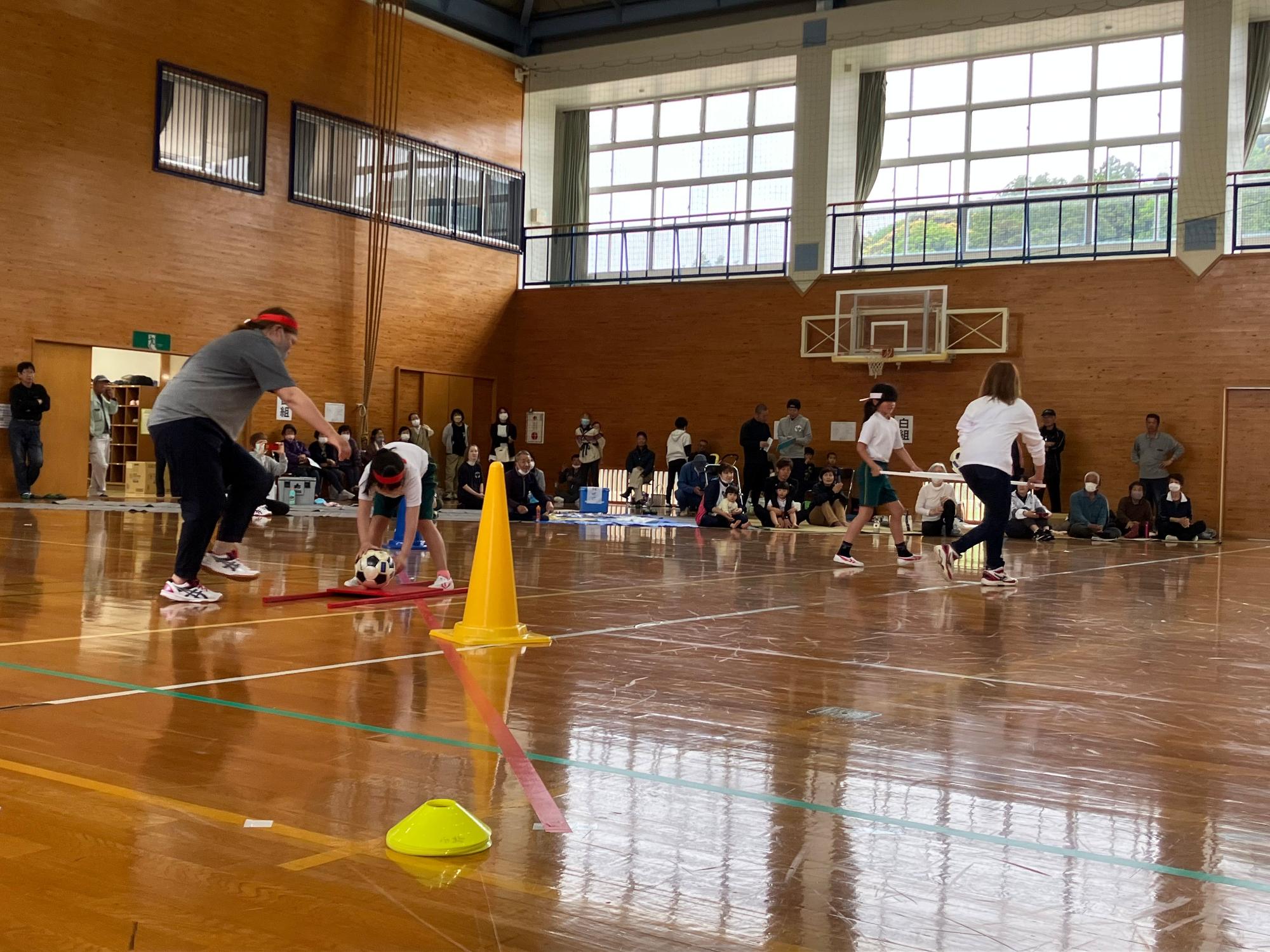 体育館の中の板張りの床の上で大人とペアになって球技に興じる児童達の様子の写真