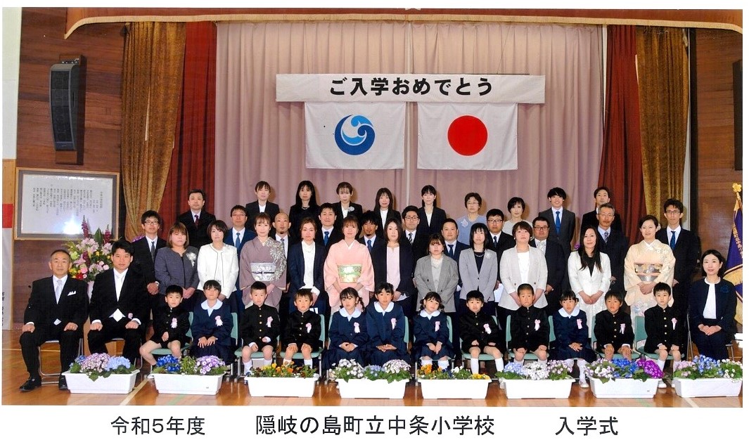 入学式で体育館の檀上に座る児童たちとその後ろに立つ保護者達の写真