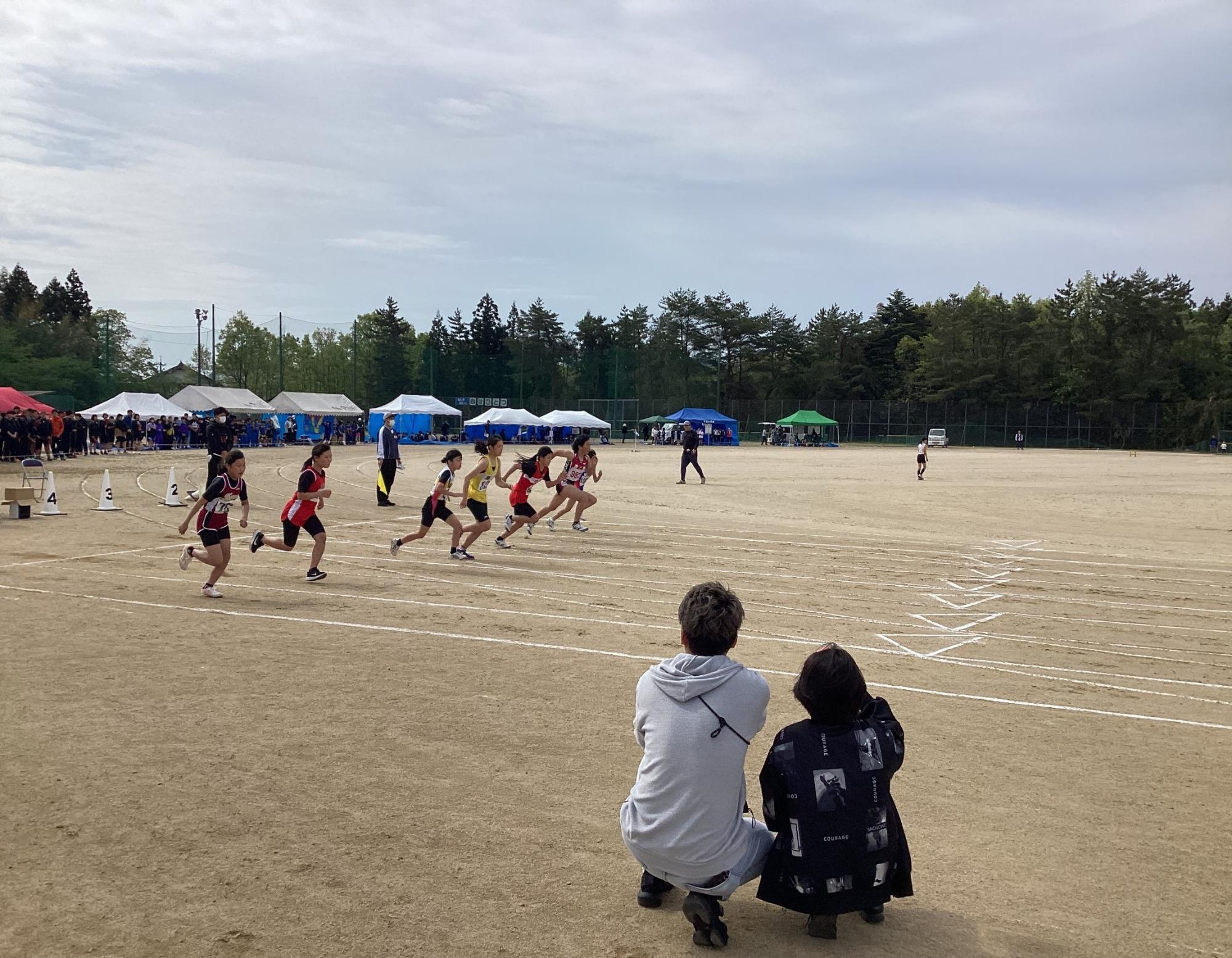 陸上大会で児童たちが一斉にスタートしたところを見守る二人の保護者の写真