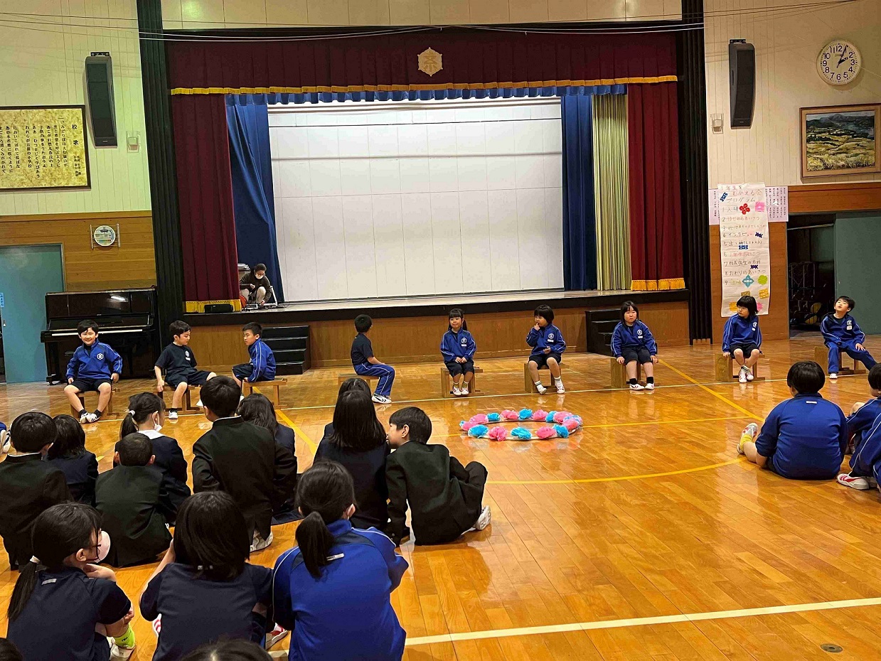 体育館の中央で円になって座る児童たちを見守る児童の写真