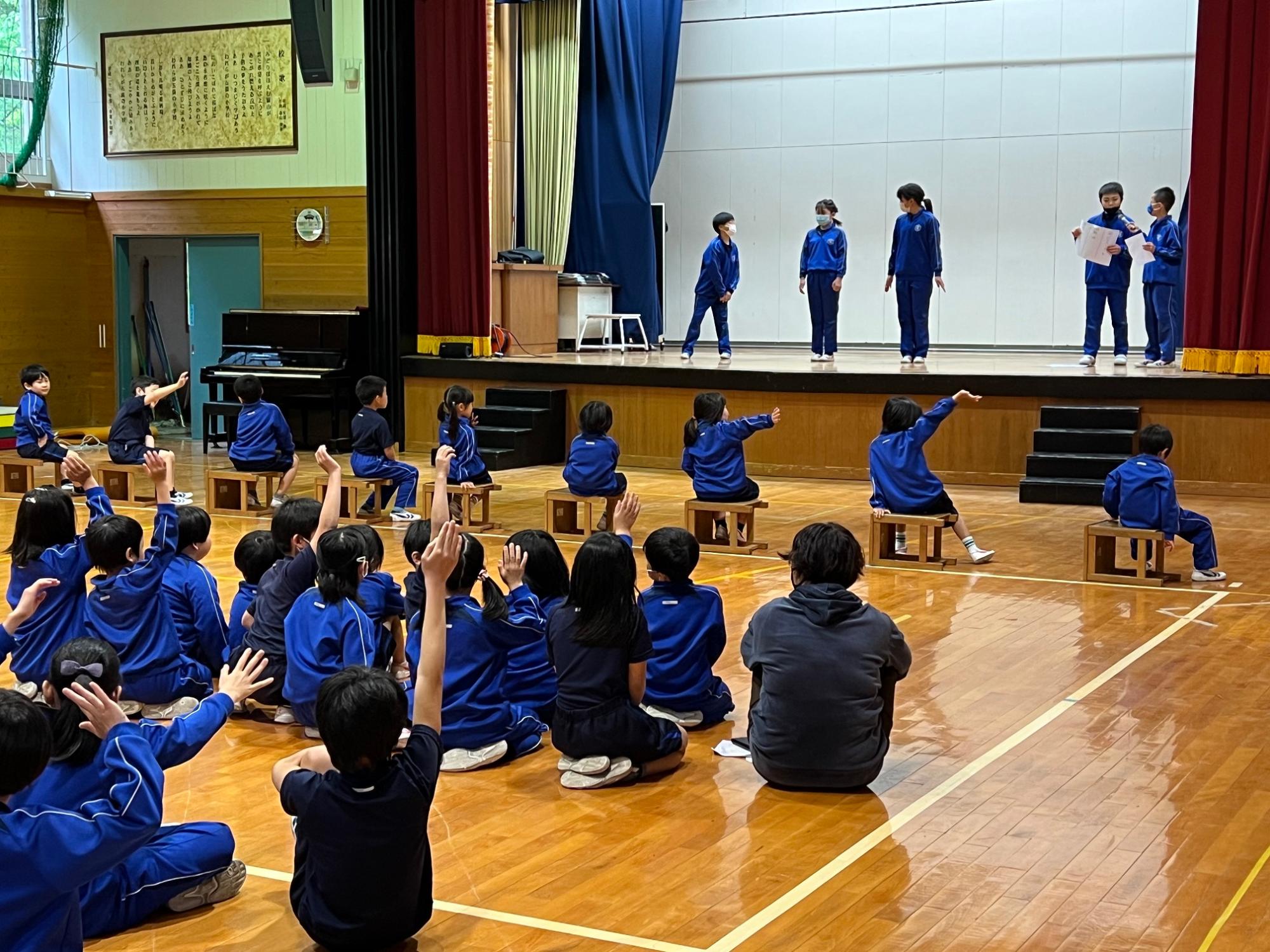 舞台上で発表する児童とそれを見て手を挙げる児童たちの写真