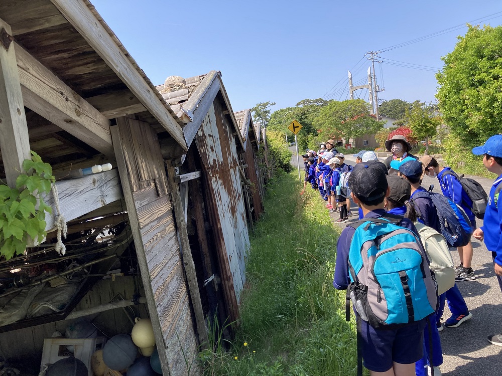 傾いた木造小屋の前で児童たちが列になって歩いている写真
