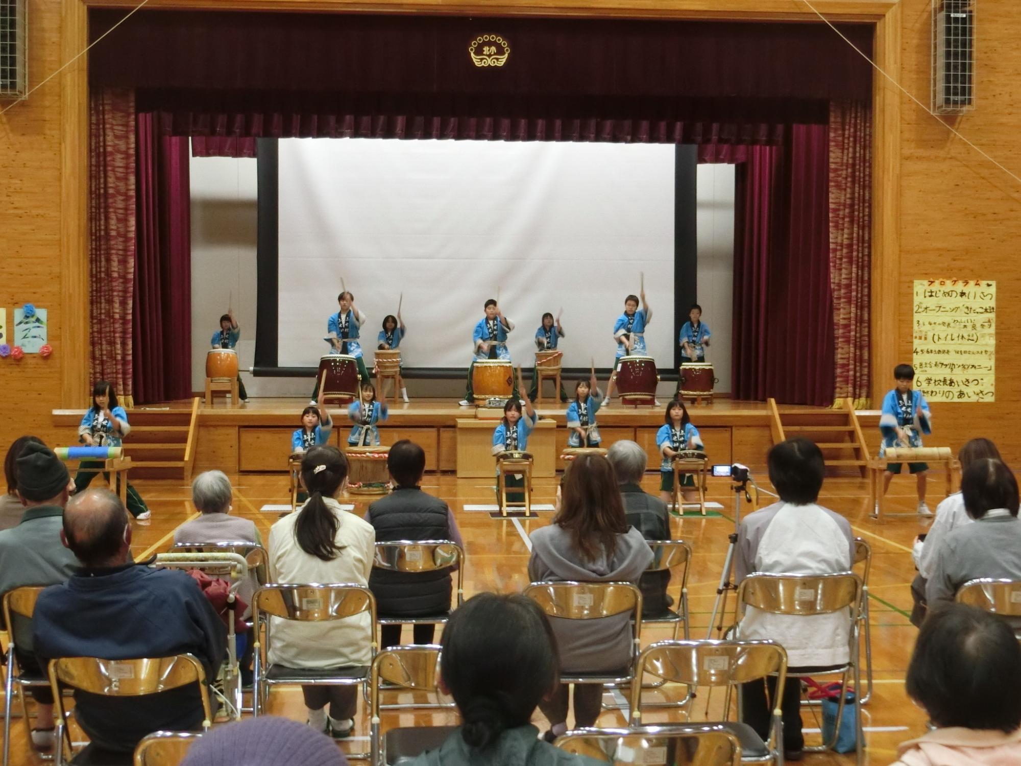 体育館の壇上でお揃いの法被を羽織ってバチを手に大きな太鼓を叩く児童たちと、パイプ椅子に腰掛けてそれを見守る大人達の様子の写真