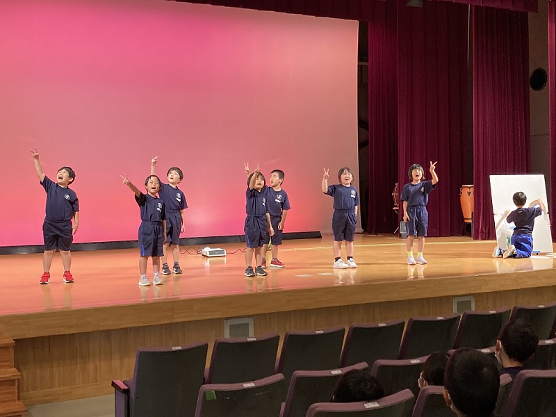 ホールの舞台で児童たちが歌のパフォーマンスを披露している様子の写真