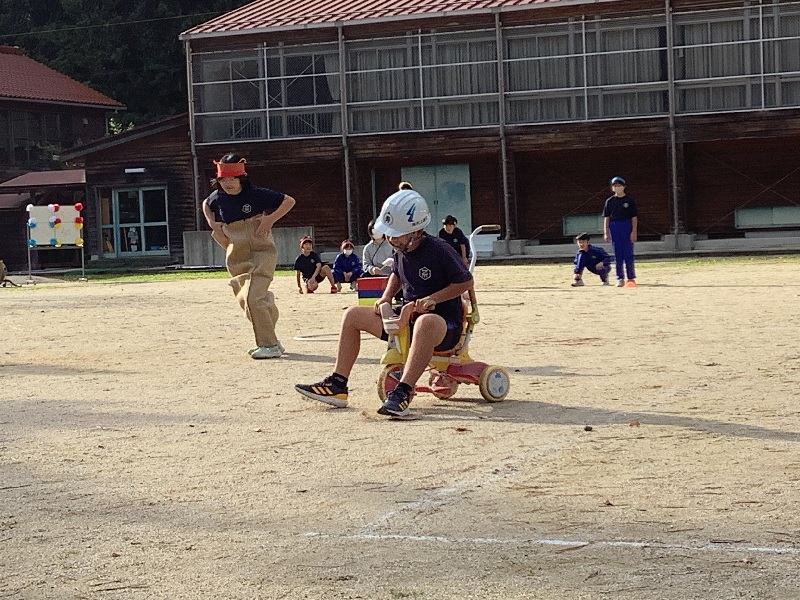 校庭でヘルメットをかぶった児童が三輪車に乗って競技をしている様子の写真