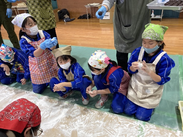 体育館で児童たちがエプロンと三角巾をつけて餅を丸めている様子の写真