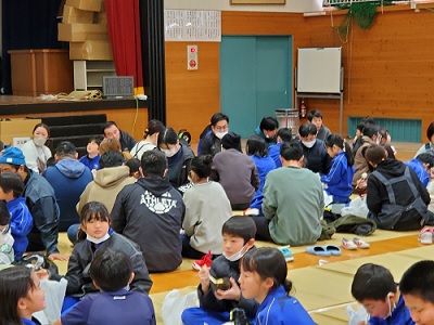 体育館で児童と保護者が円になって座り、交流しながら餅を食べている様子の写真