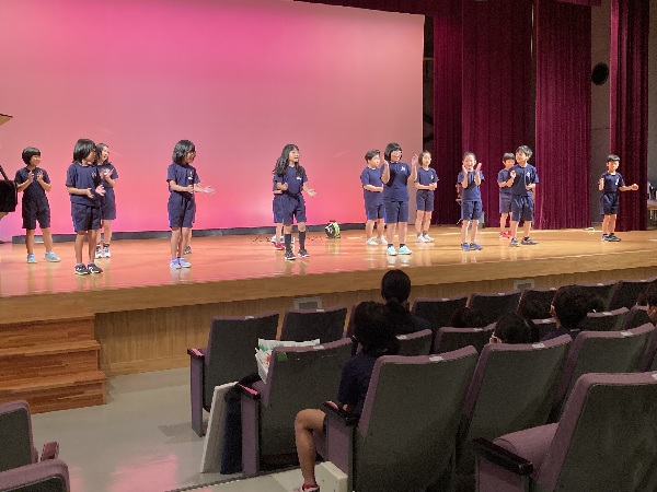 ホールの舞台で児童たちが手拍子をしながら歌っている様子の写真