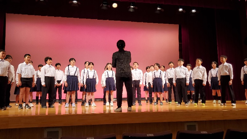 ホールの舞台で児童たちが指揮に合わせて合唱している様子の写真