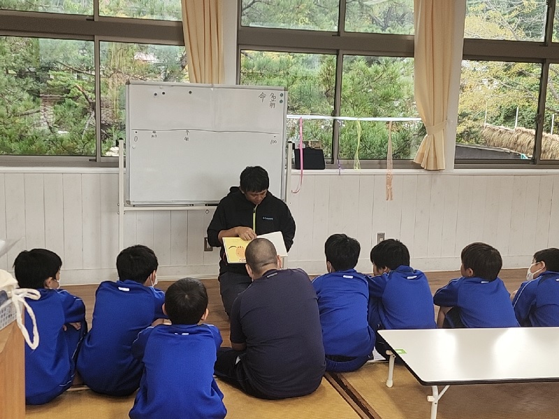 教室で大人が絵本を読み、児童たちが囲んで聞いている様子の写真