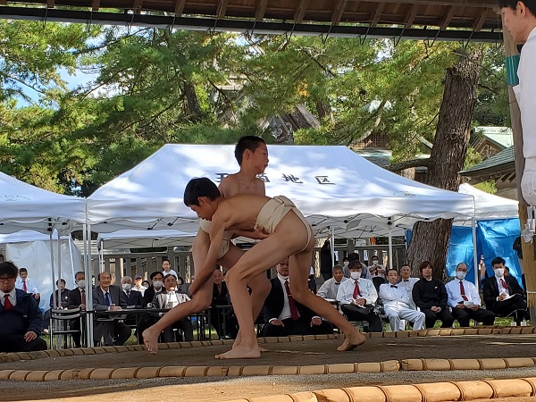 屋外の土俵で児童たちが観客に見守られながら相撲を取っている様子の写真