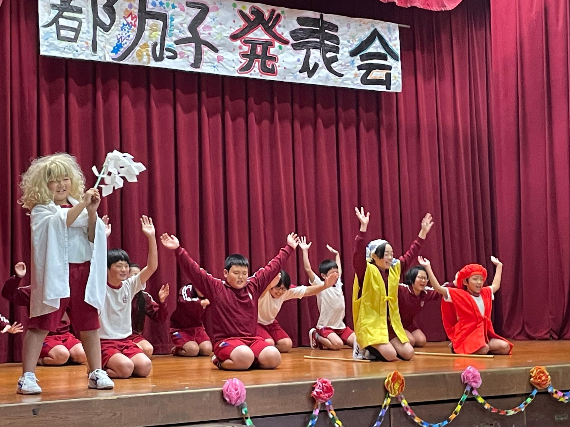 ステージで赤い服を着た児童たちが手を挙げて演技している写真