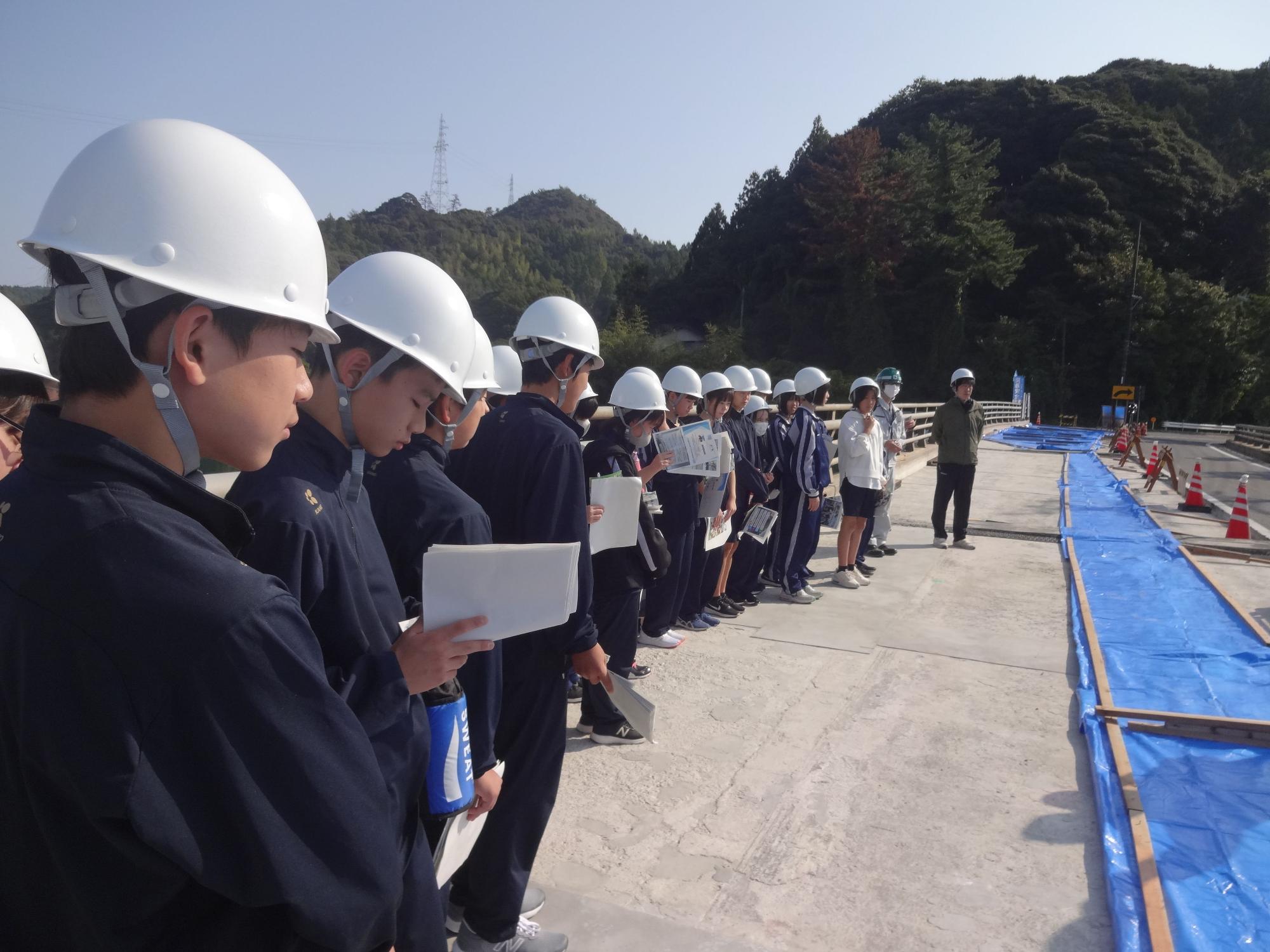 工事現場の見学でヘルメットをかぶった生徒たちが説明を聞いている写真