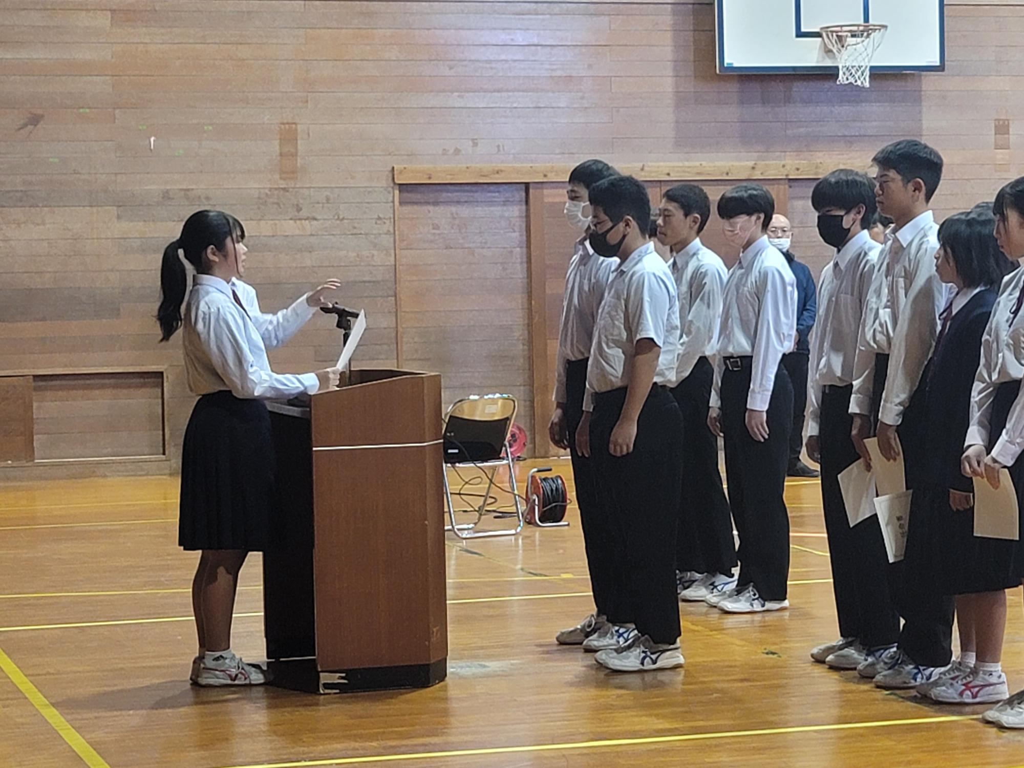 体育館でクラスの生徒たちが女子生徒の演説を聞いている写真
