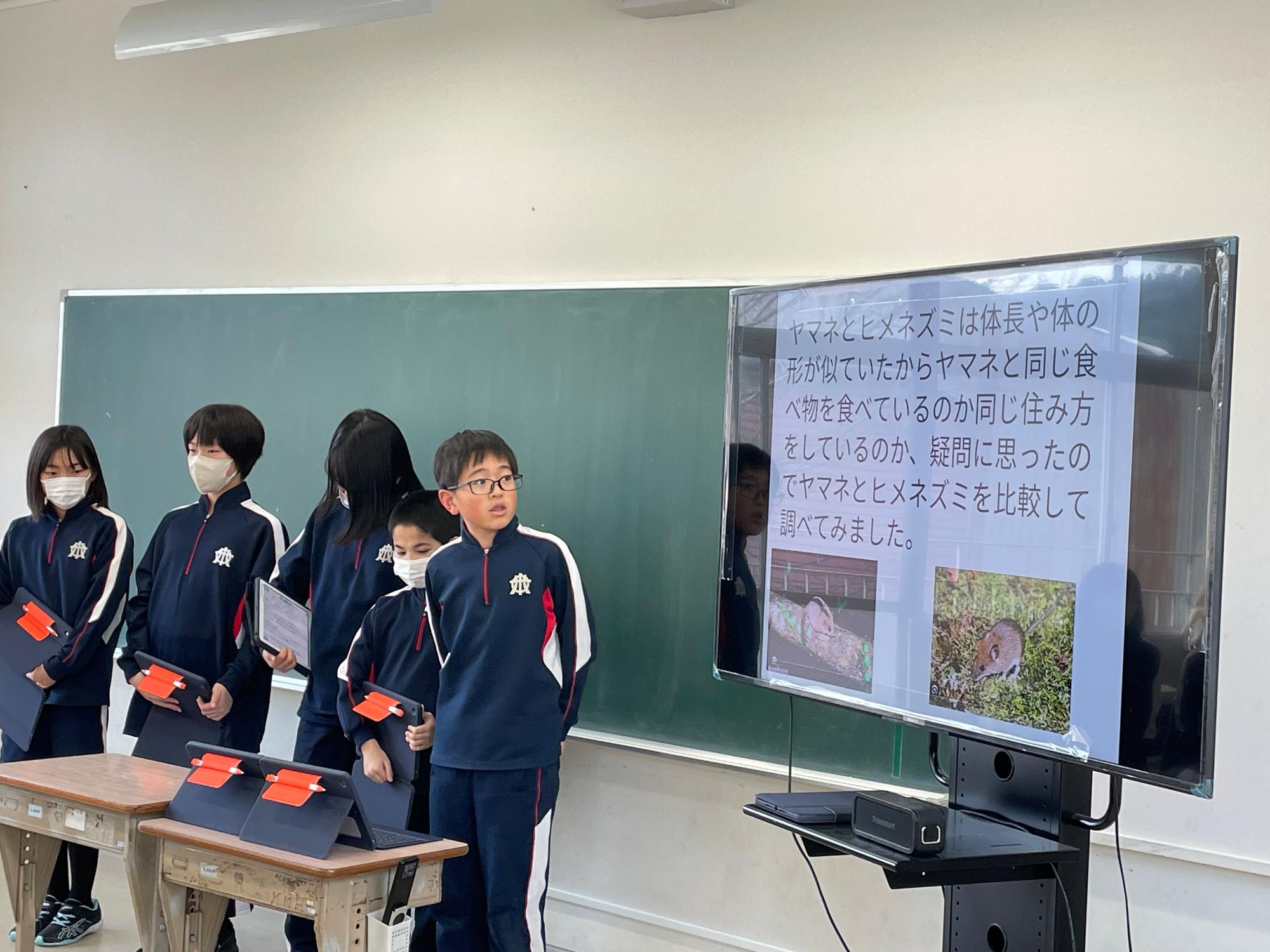 教室の黒板の前で児童たちが大型モニターとタブレットを使って発表している写真