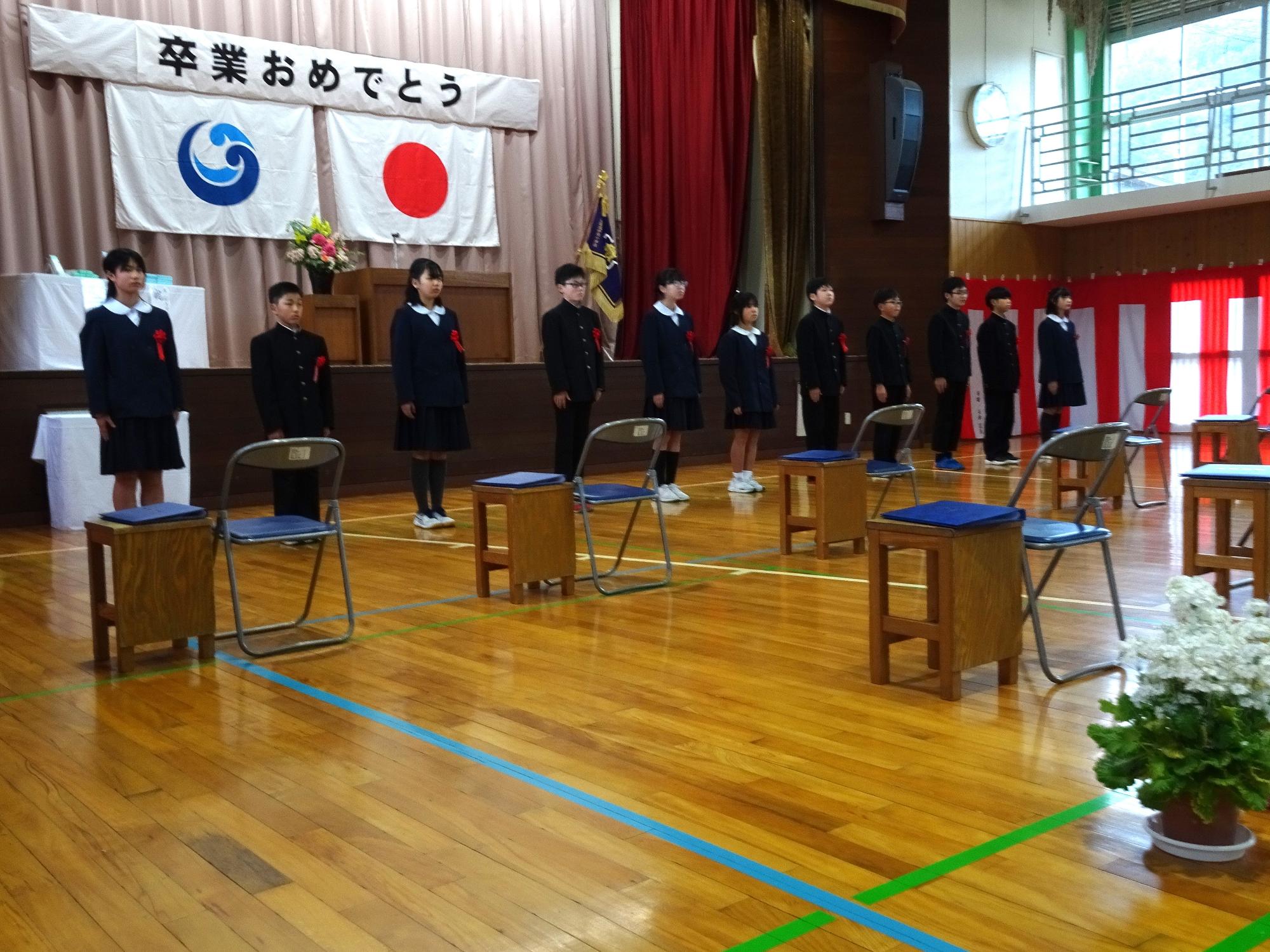 体育館で卒業生の児童たちが卒業式に臨み、国旗の掲げられた壇上を背に横一列で整列している写真