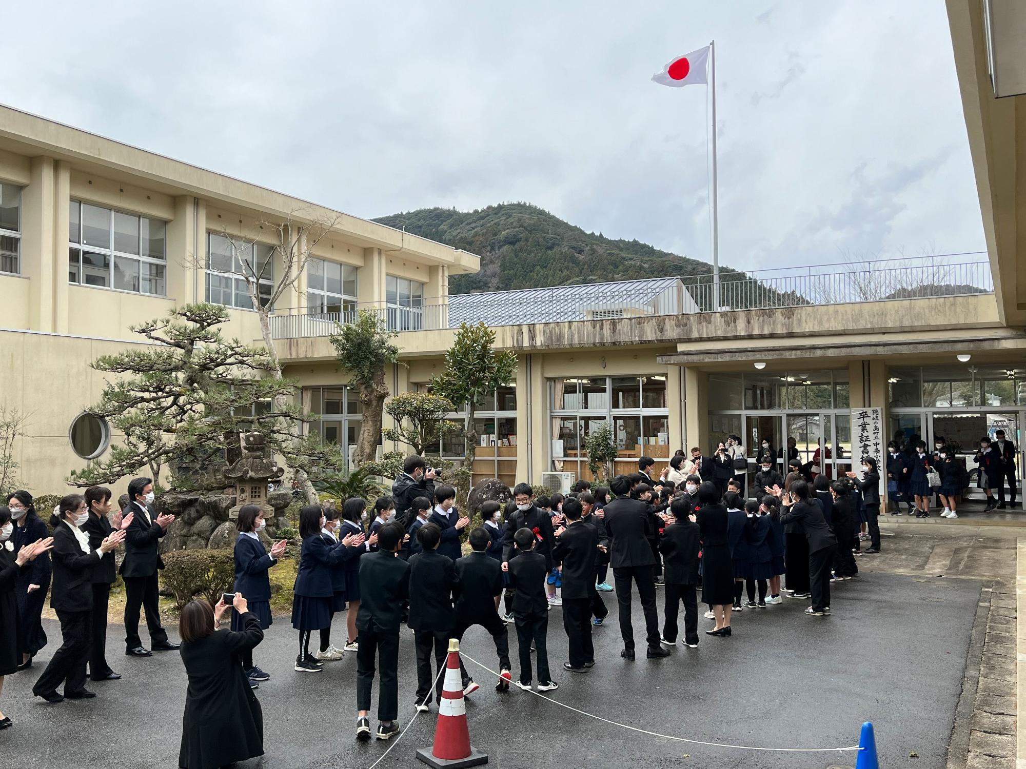 校舎前で在校生や保護者たちが花道をつくり卒業生を拍手で見送っている写真