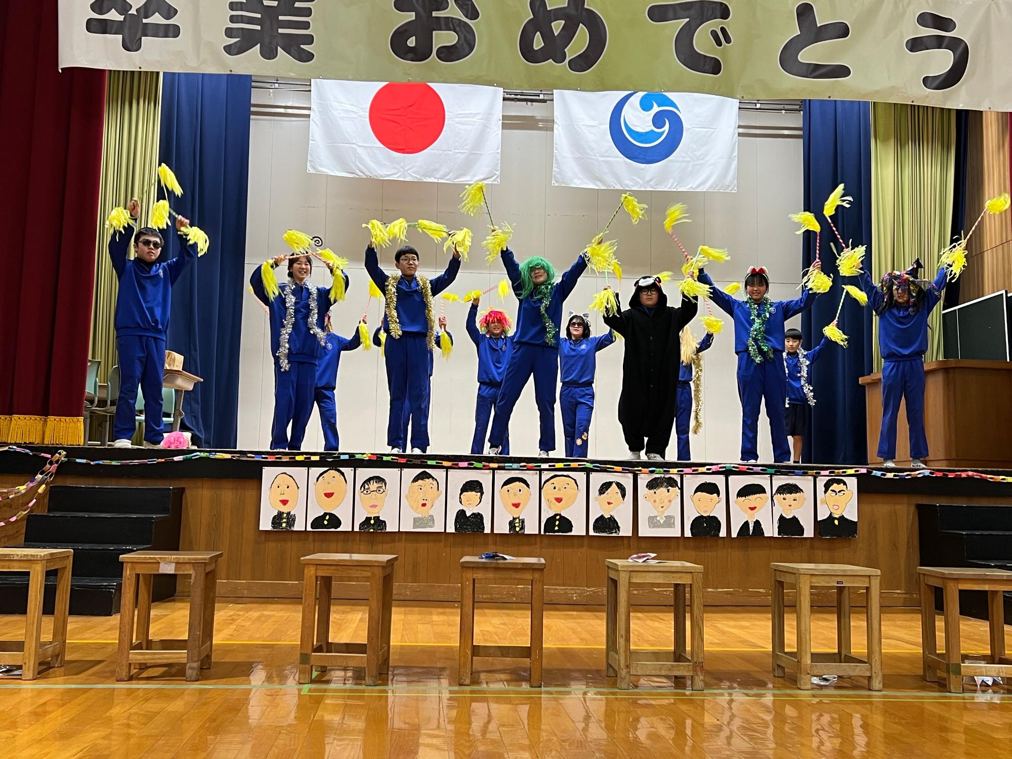 卒業おめでとうの垂れ幕の下で被り物をした児童たちが黄色のポンポンを持ってダンスをしている写真