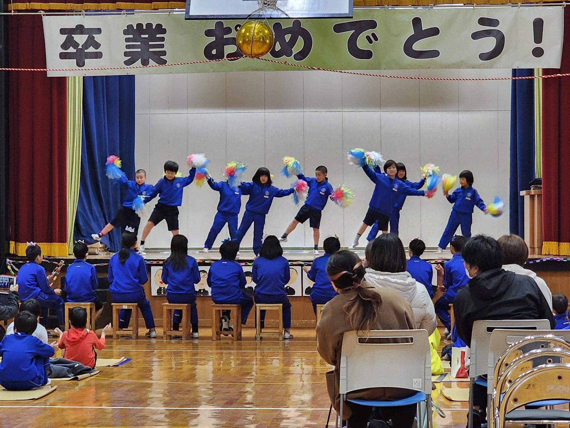 舞台上で児童たちがカラフルな色のポンポンを持ってダンスを披露している様子を椅子に座った児童と先生が見ている写真