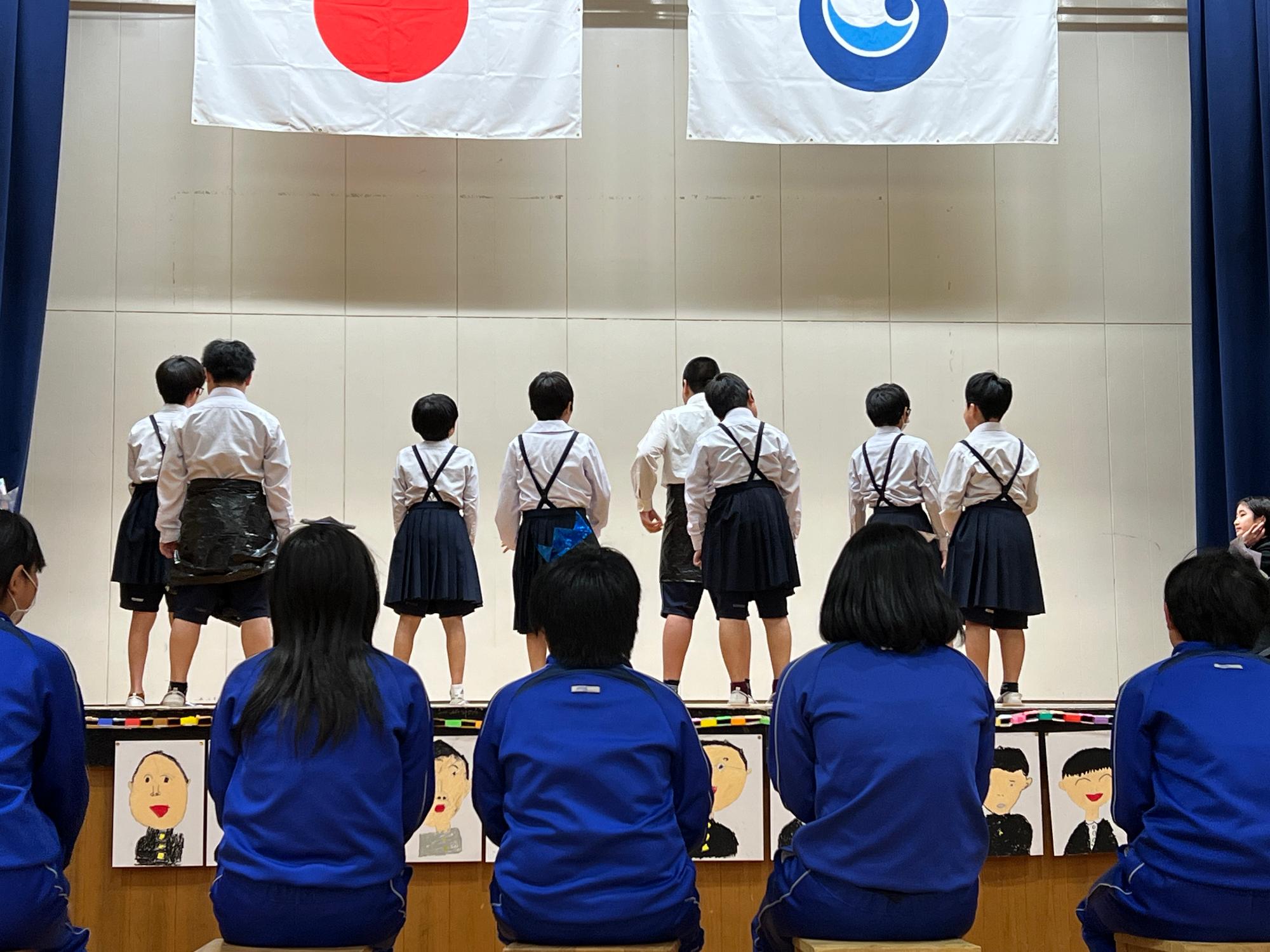 体育館の舞台で制服姿の児童たちが背中を向けて立っている写真