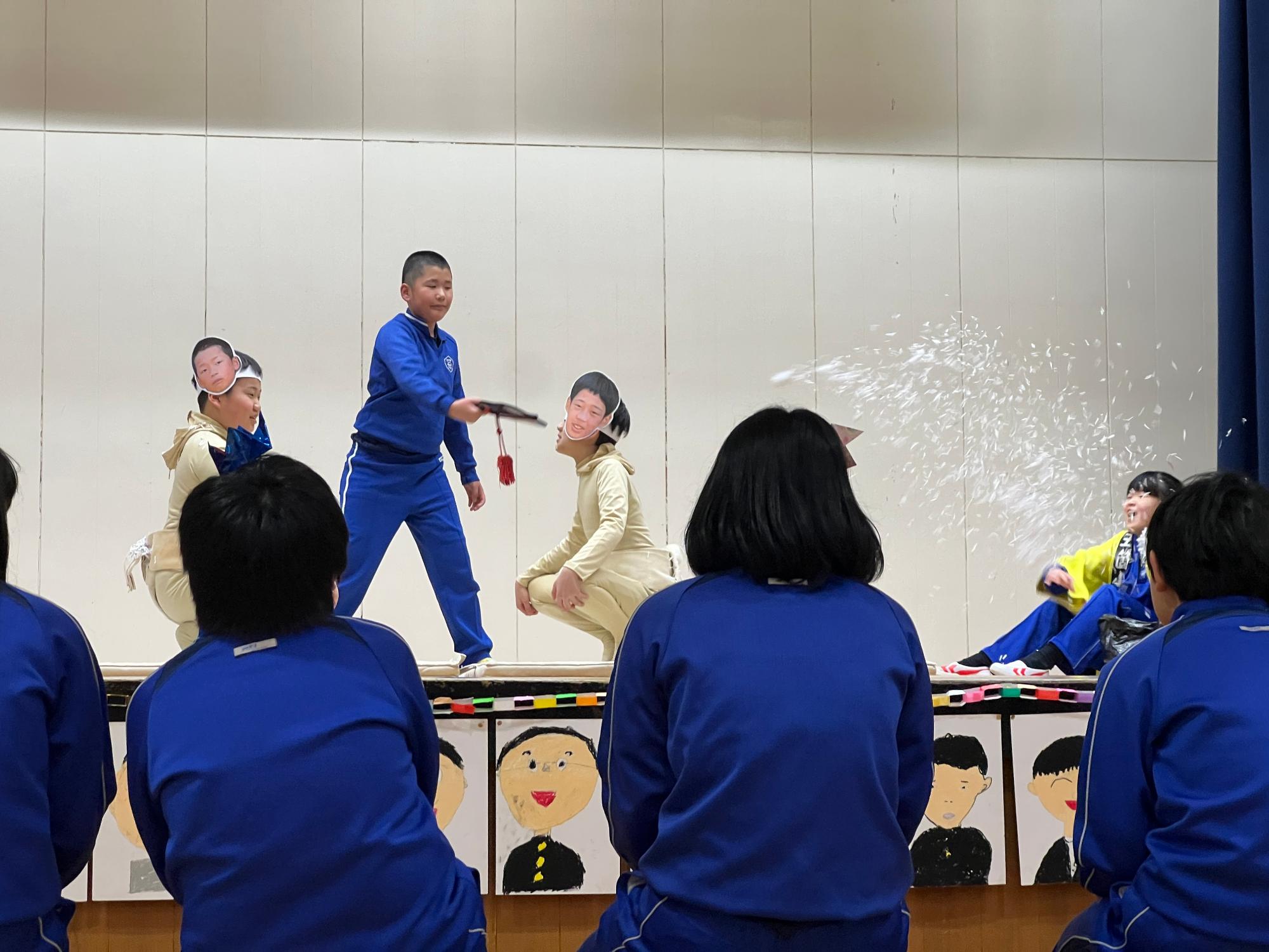 体育館の舞台で児童が顔写真の仮面をつけて劇をしている写真