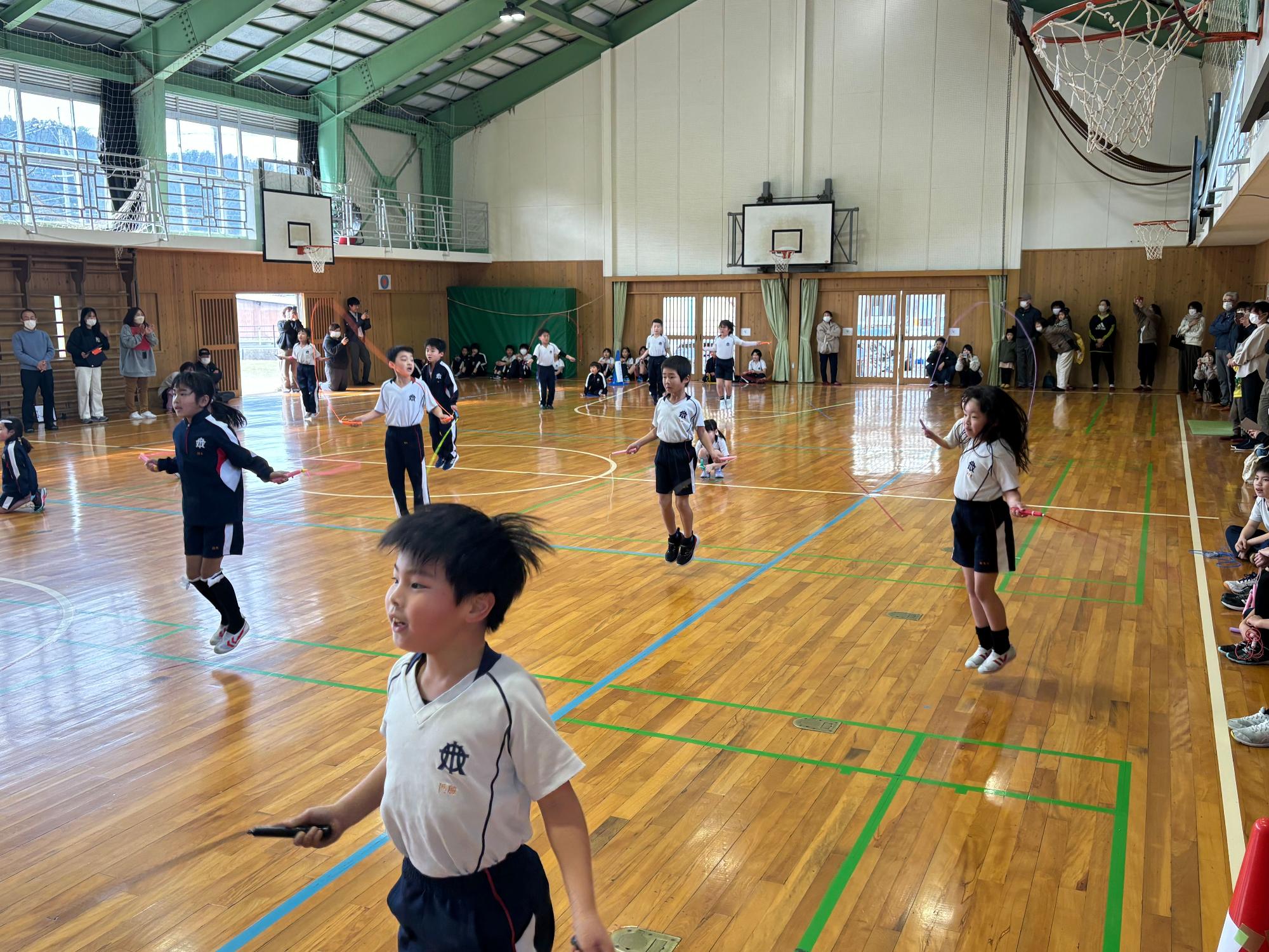 体育館で児童たちが観客に見守られながらなわとびに取り組む様子の写真