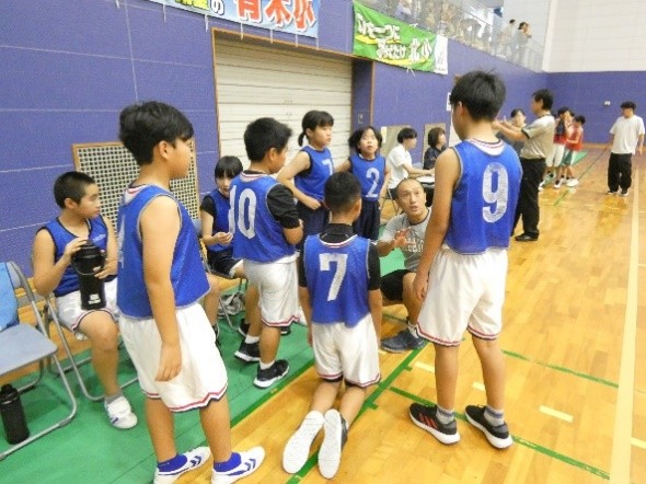 体育館で青いビブスを着た児童たちがコーチの話を聞いているバスケットボールチームの写真
