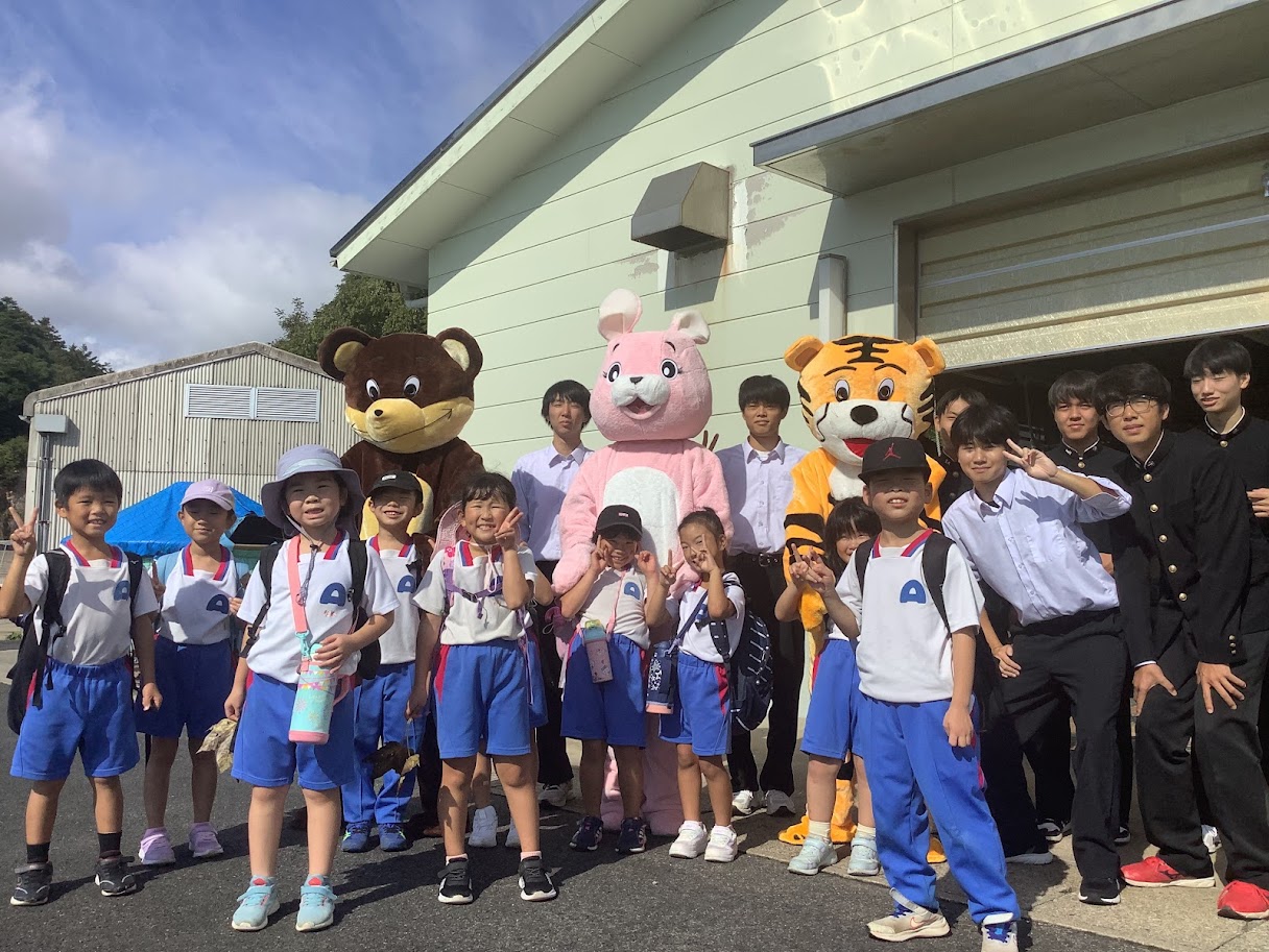 着ぐるみを着たキャラクターと小学生・高校生たちが外で記念撮影している写真