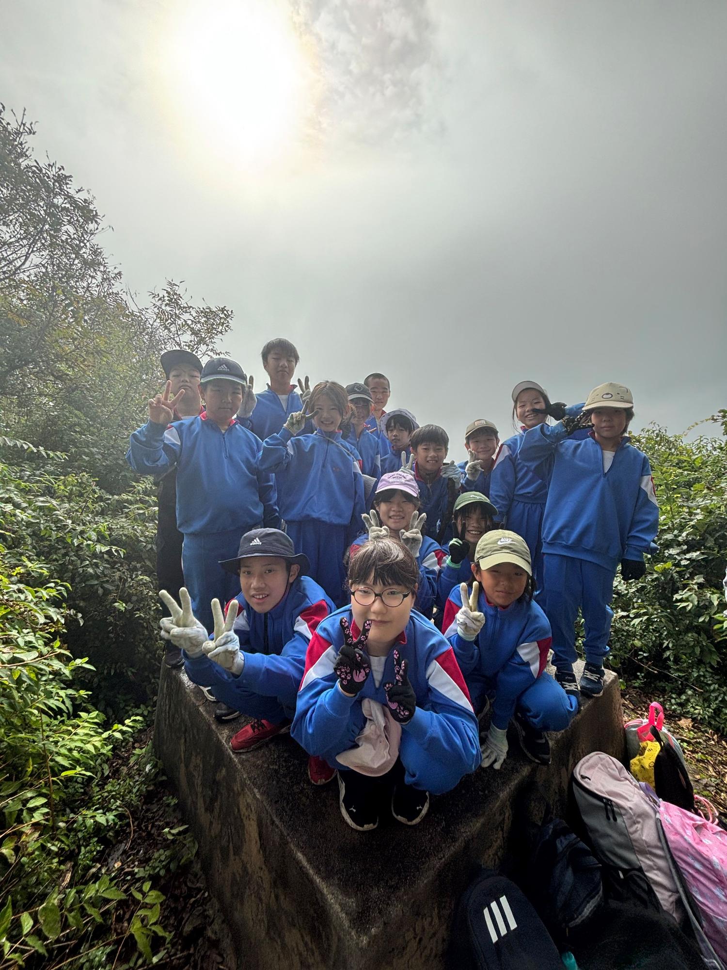 山頂付近で青いジャージ姿の子どもたちが集合しピースをしている登山の記念写真