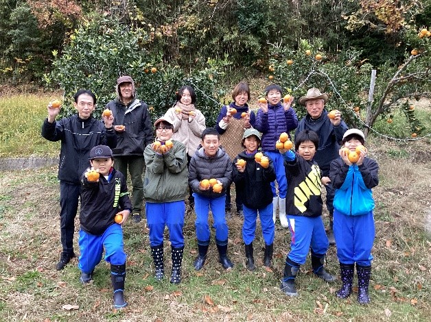 みかんの木の前で子どもと大人がみかんを手に持って笑顔で集合している写真