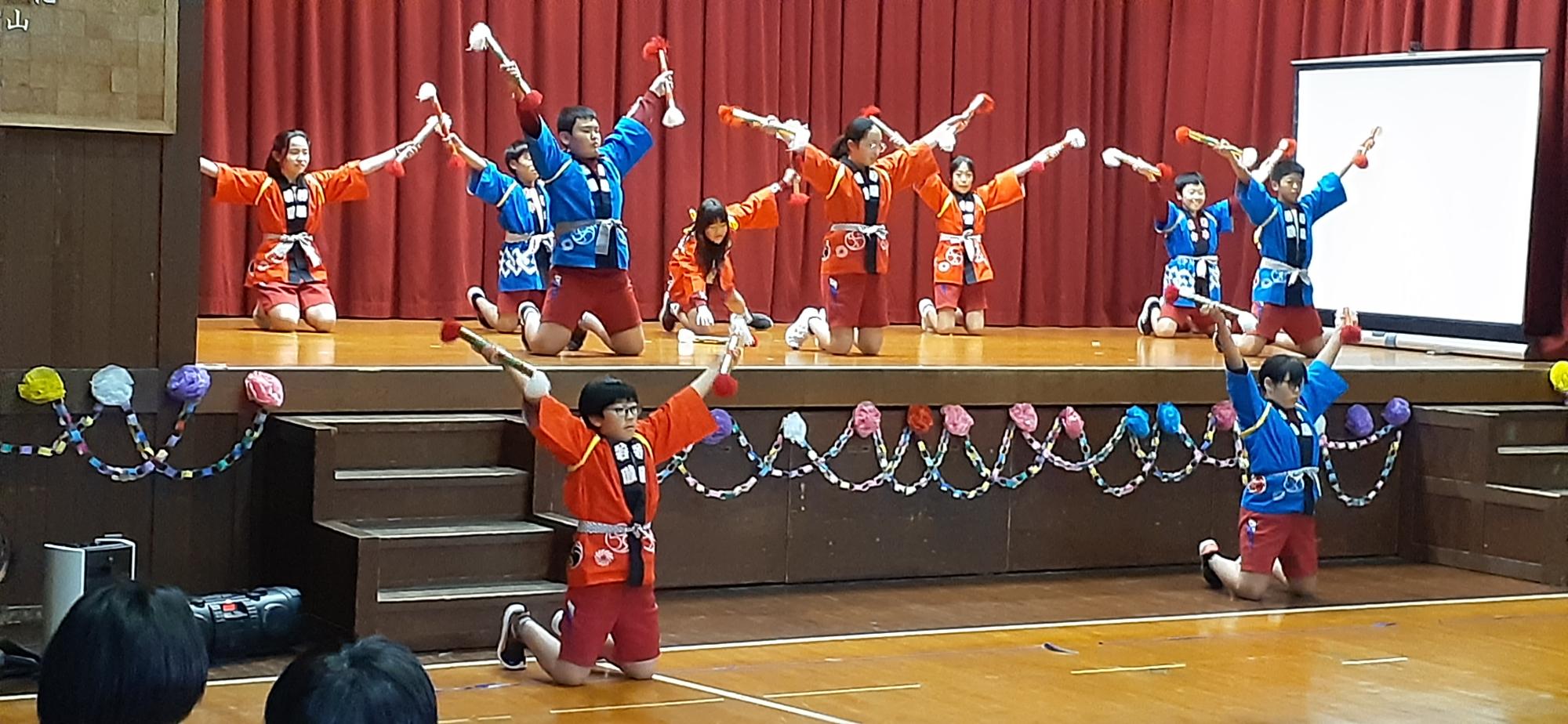体育館の舞台で児童たちが法被を着て踊りを披露している写真