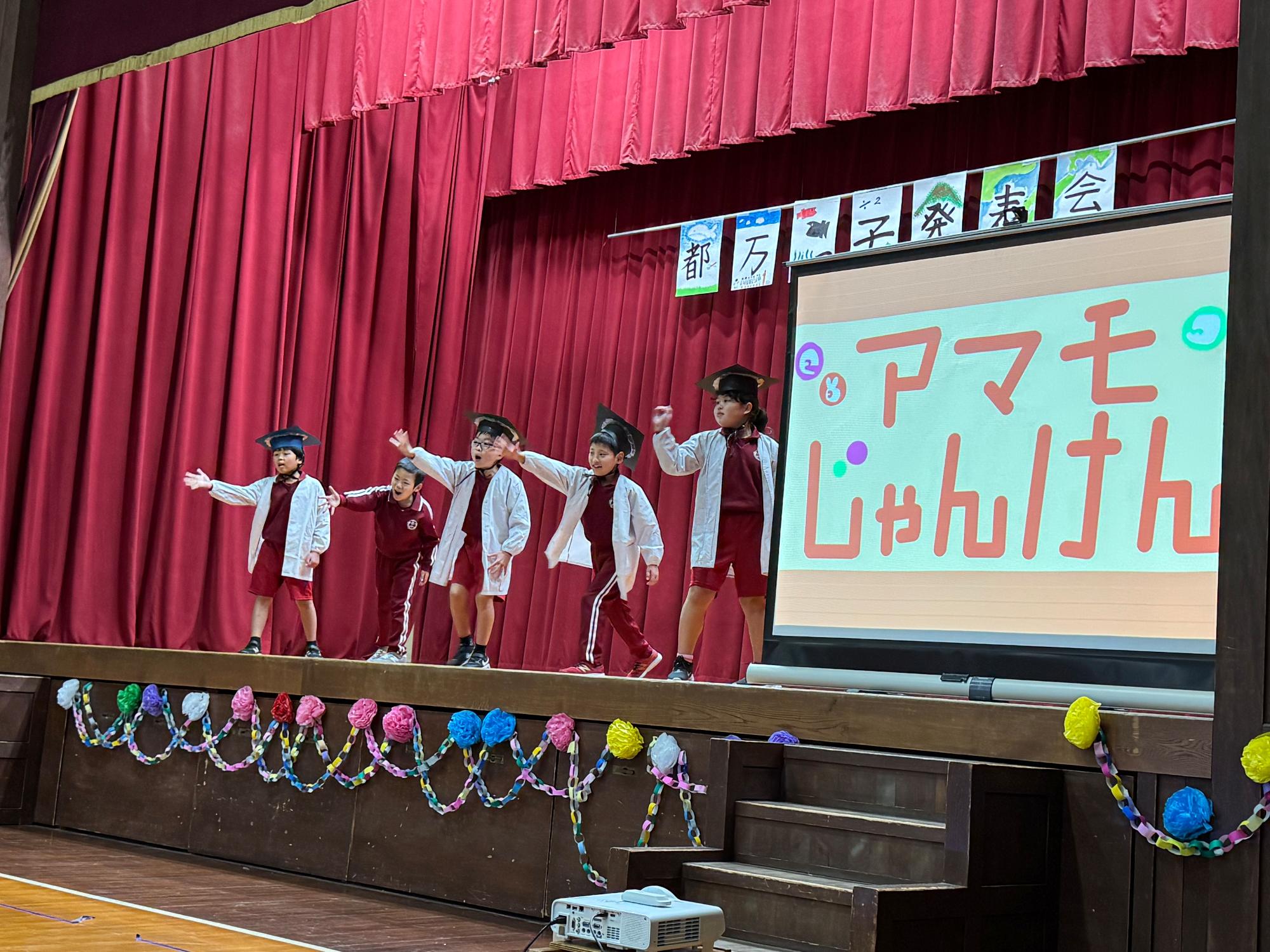 体育館の舞台で児童たちがアマモじゃんけんの発表をしている写真