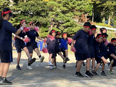 外で児童たちが肩を組みダンスを踊っている写真