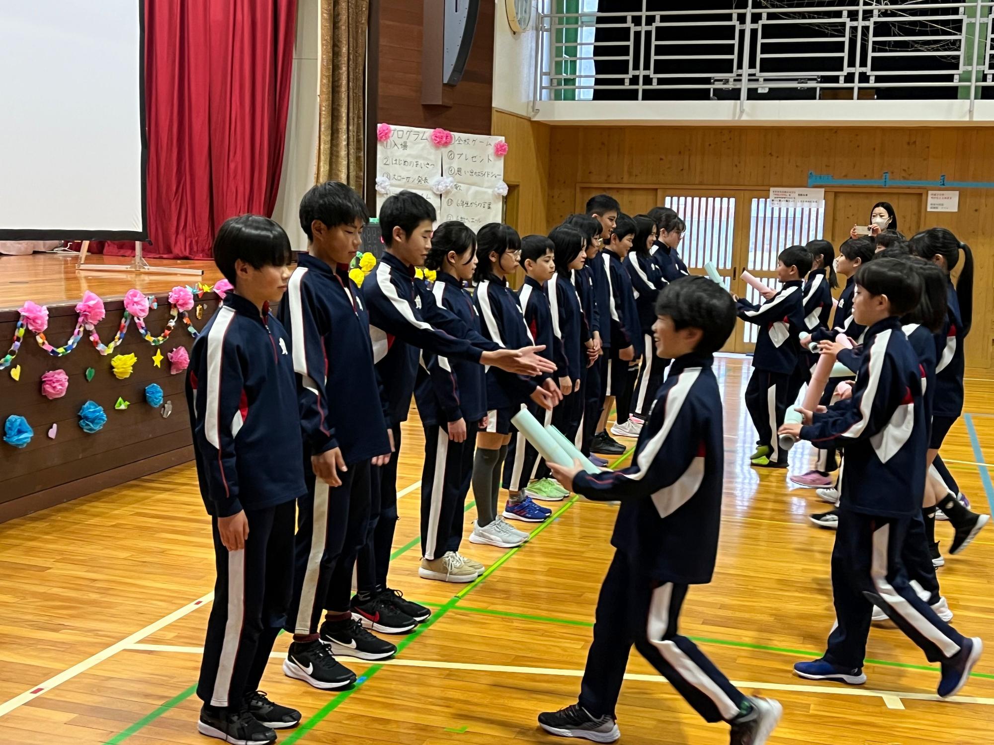 体育館で児童たちが先輩に贈り物を渡している写真