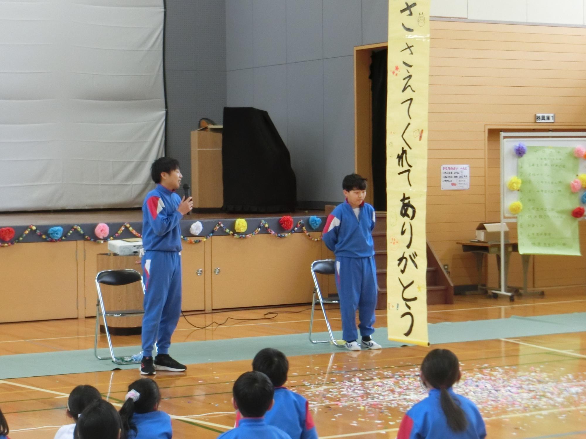 体育館で座っている六年生に向けてマイクを持った在校生がメッセージを伝えている写真