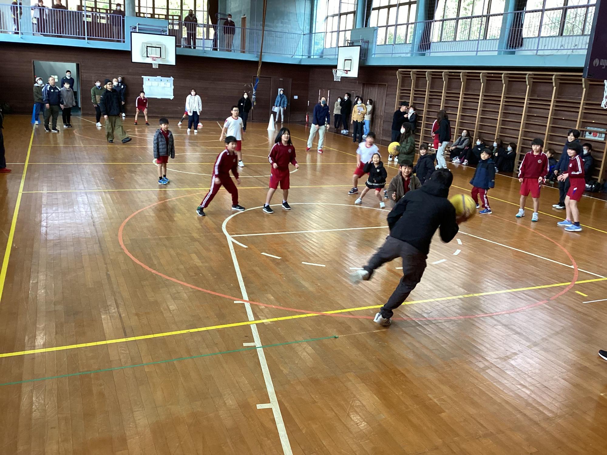 体育館で体育着を着た児童たちがドッジボールで遊び、その様子を近く見守る大人たちの写真