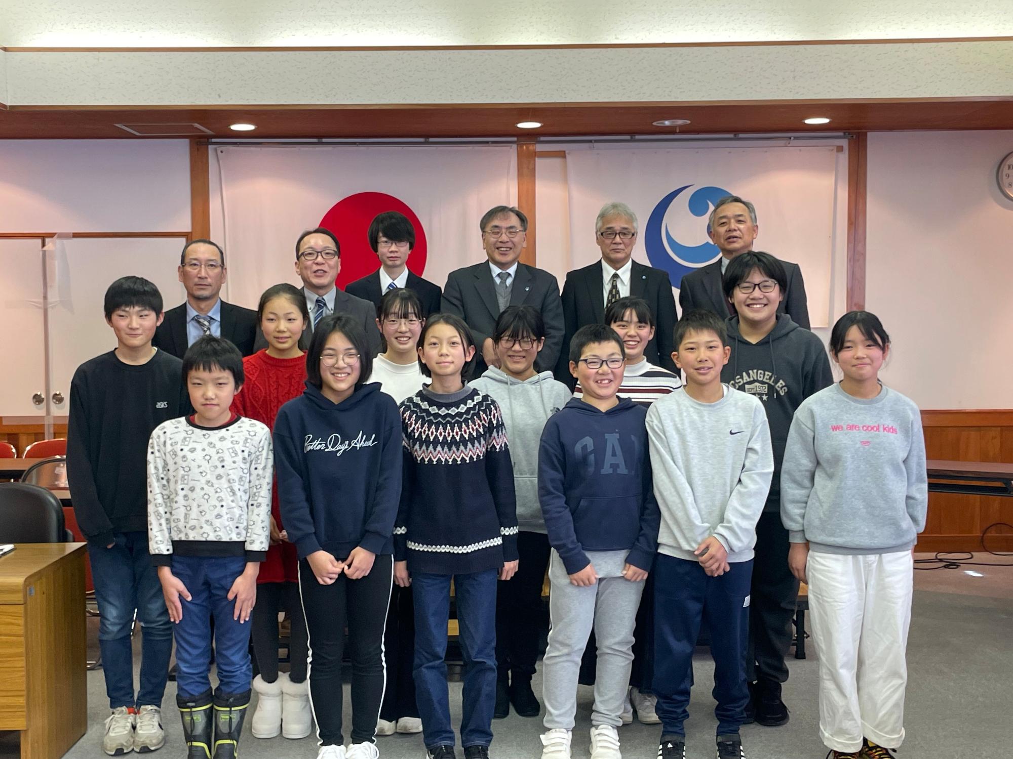 会議室で複数人の児童とスーツを着た大人たちが並んで記念撮影をしている写真