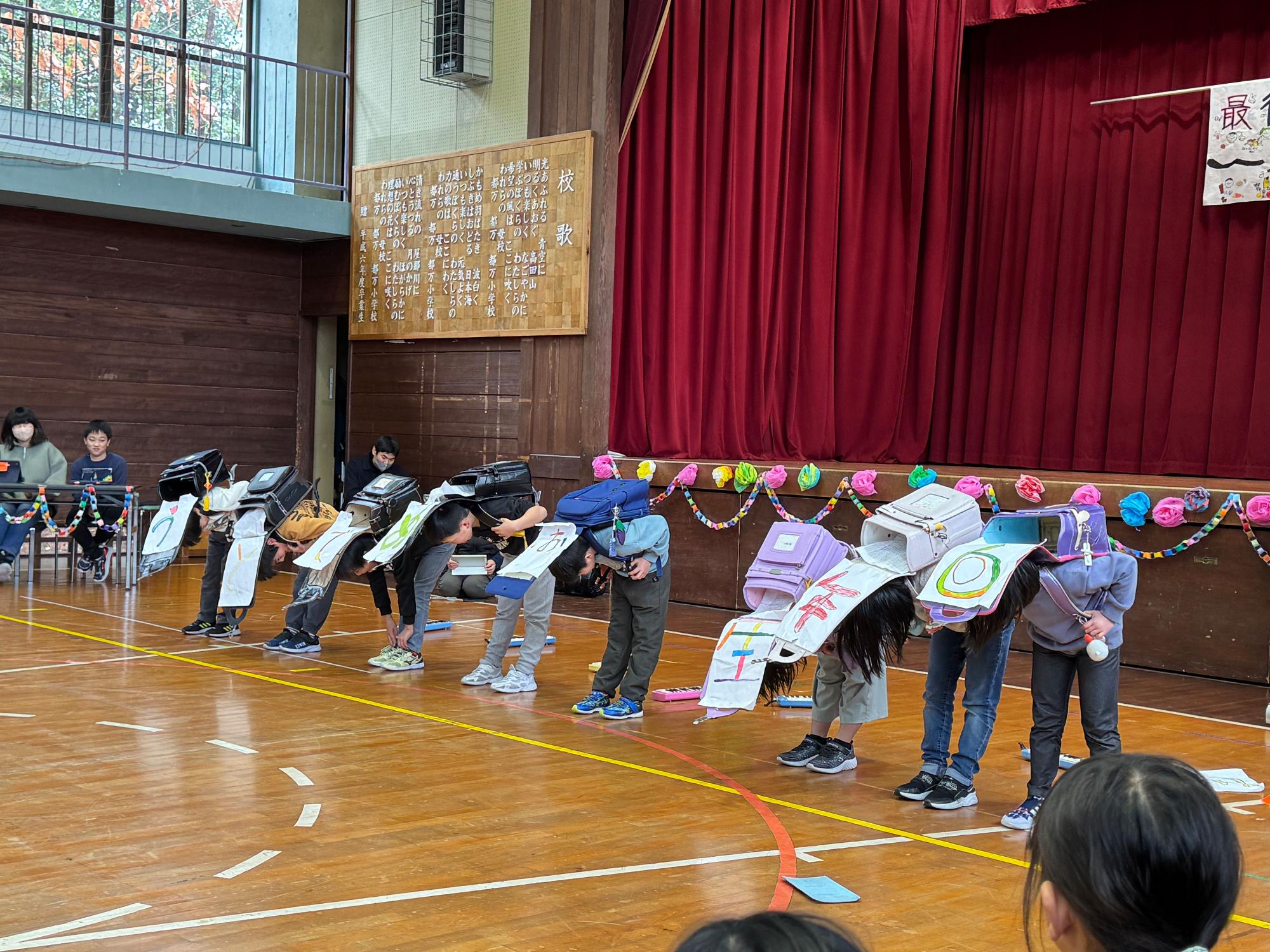 体育館で児童たちが一列に並びランドセルを背負っておじぎをしている写真