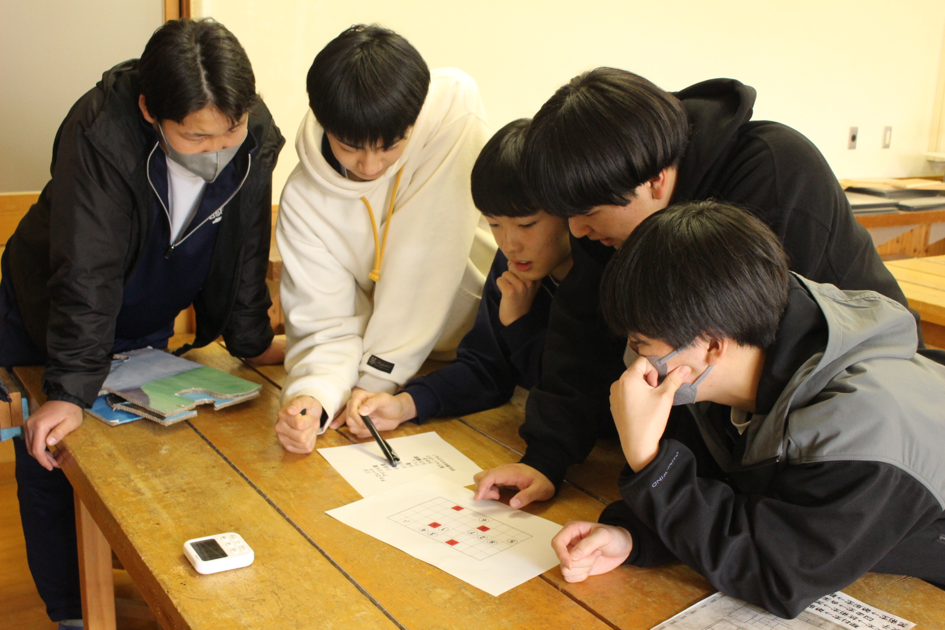 教室で男子生徒たちが紙を囲んで紙で遊べるゲームをしている写真