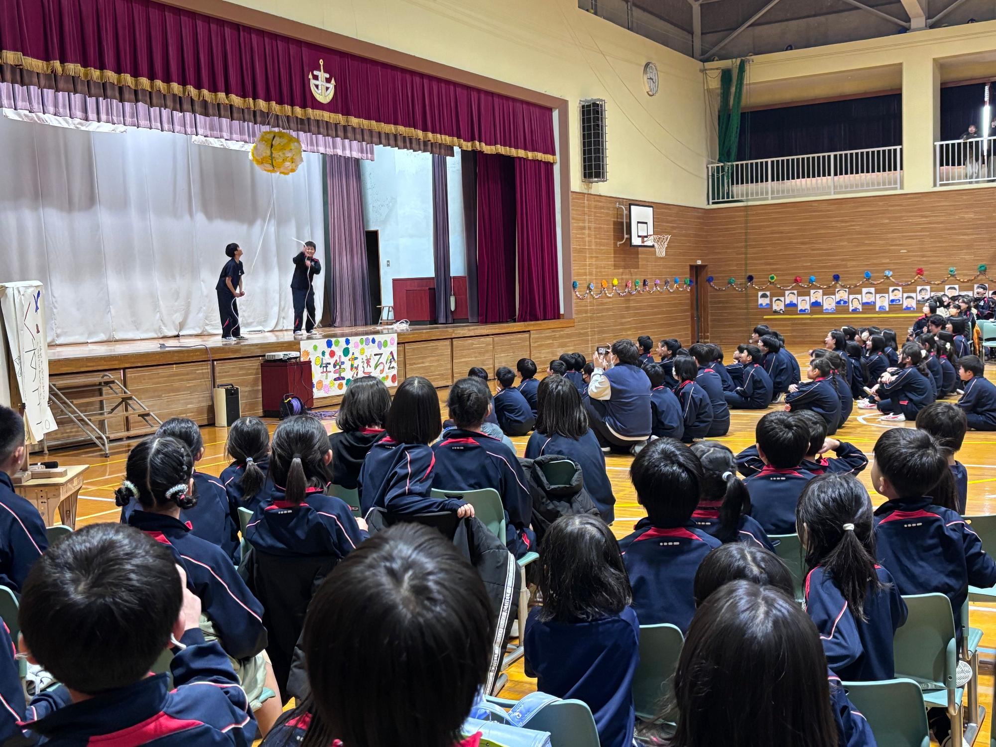 体育館で児童たちが舞台発表を見ているところを後ろから撮影した写真